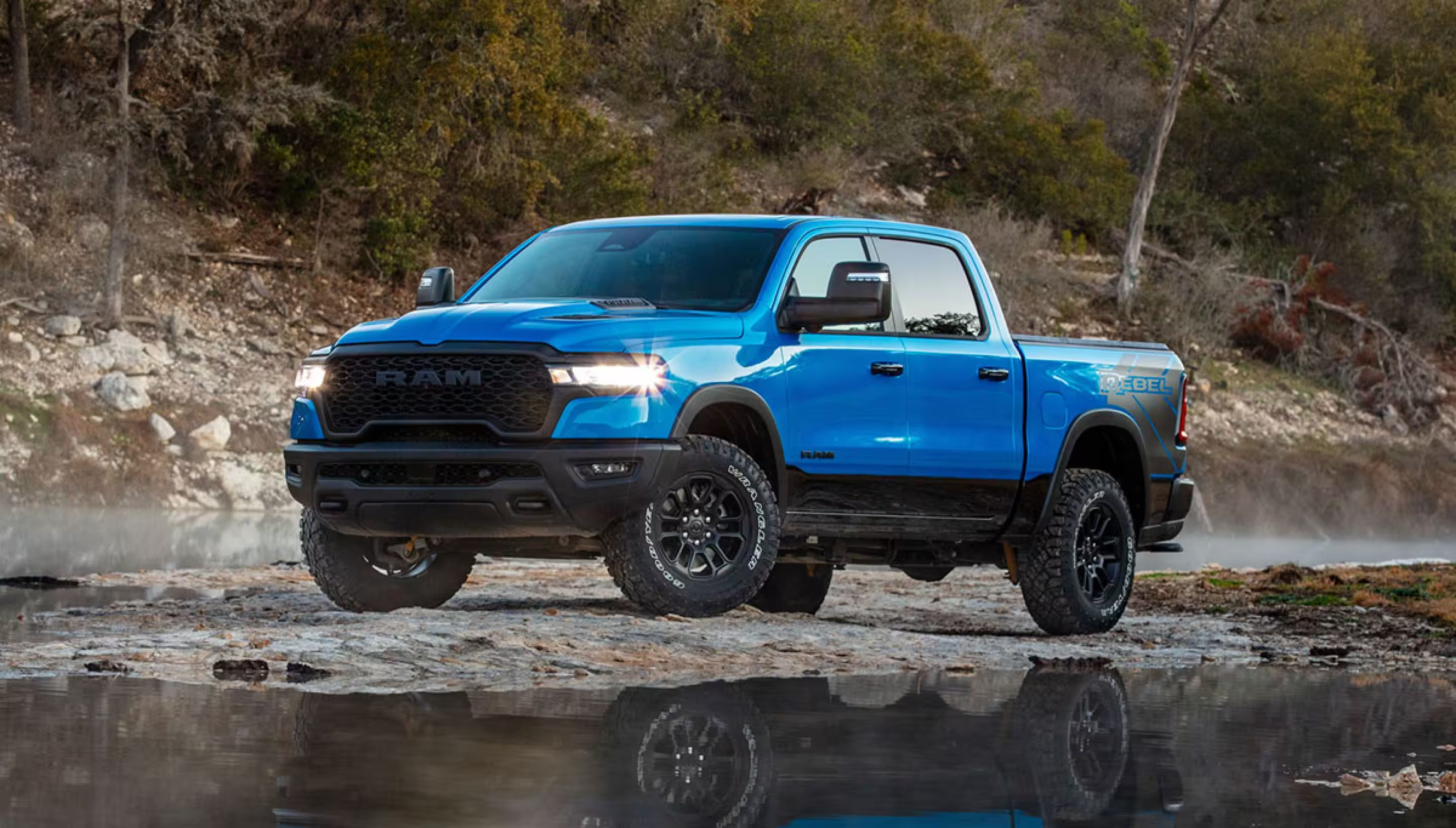A blue 2025 Ram 1500 Rebel Crew Cab being driven on a muddy trail off-road, beside a large hill.