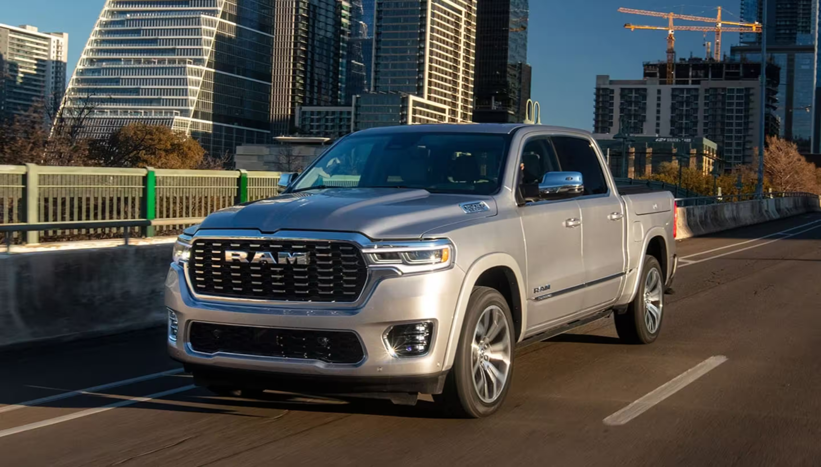 A silver 2025 Ram 1500 Tungsten Crew Cab being driven on a highway bridge in a city, with tall buildings in the background.