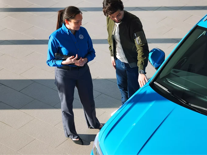 Standing beside a Taos shown in Cornflower Blue, an interested customer speaks with a Volkswagen sales/product specialist at a dealership