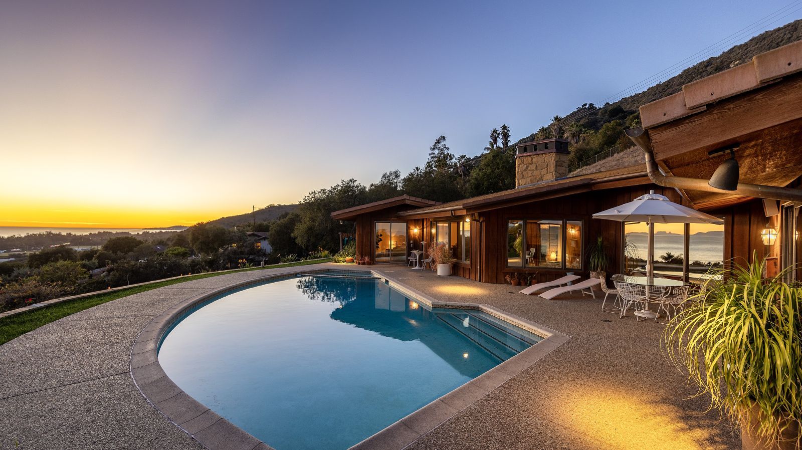 A swimming pool surrounded by a terrace adjacent to a luxury home and looking out to a distant ocean view at sunset.