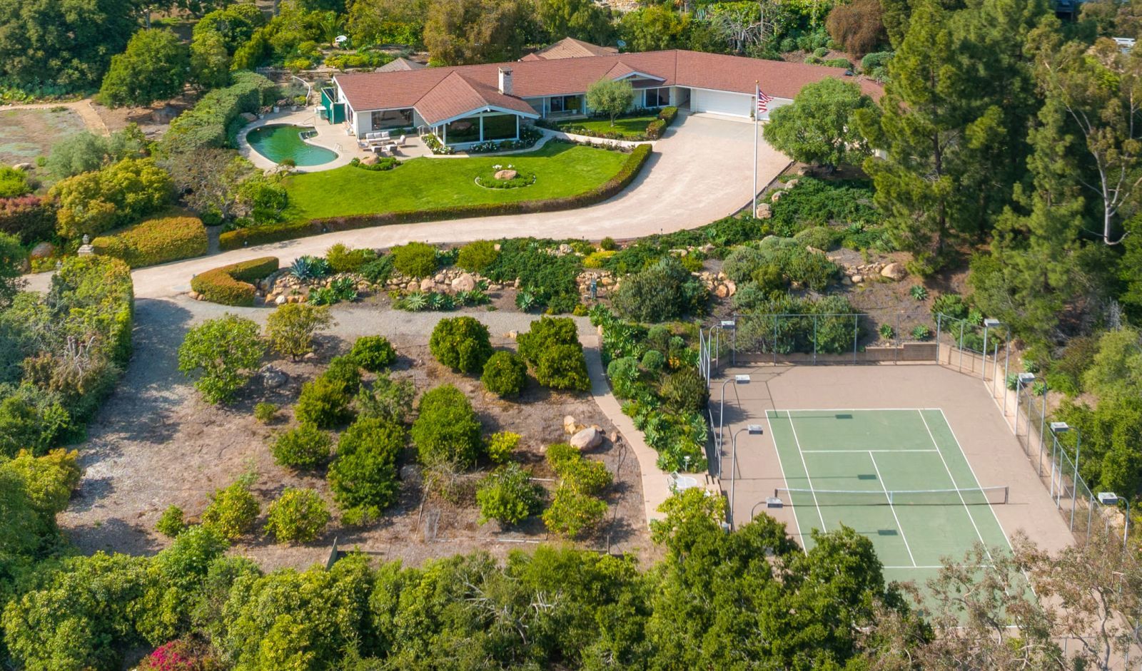 A birds eye view of a California estate with a winding road from the tennis court to the ranch-style house surrounded by manicured landscaping