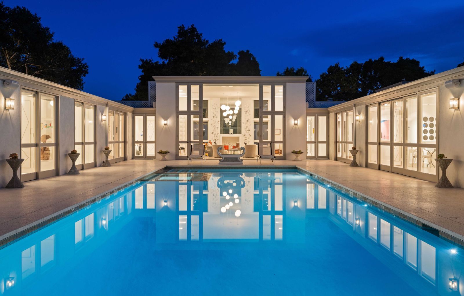 The pool courtyard of a large mid-century modern home in Montecito, the expansive pool flanked by rows of illuminated floor-to-ceiling glass windows and doors.