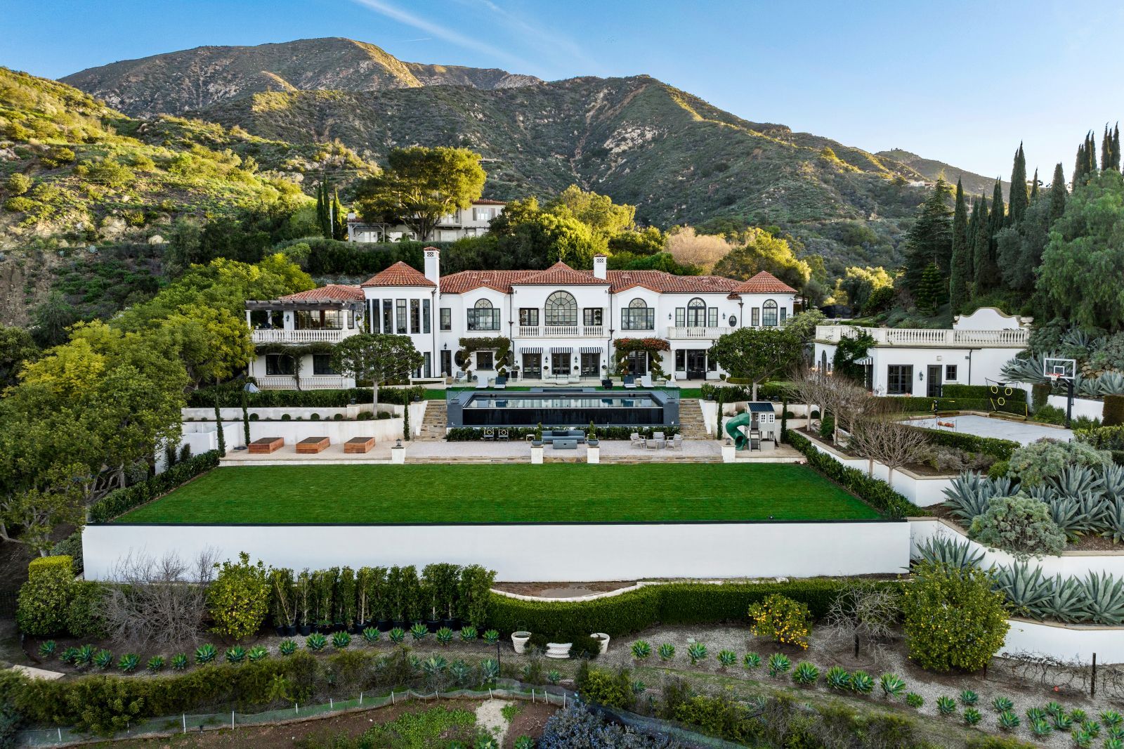 A Montecito estate with its large red tiled roof Mediterranean-style home and mountains in the background and lush lawn and blue swimming pool.