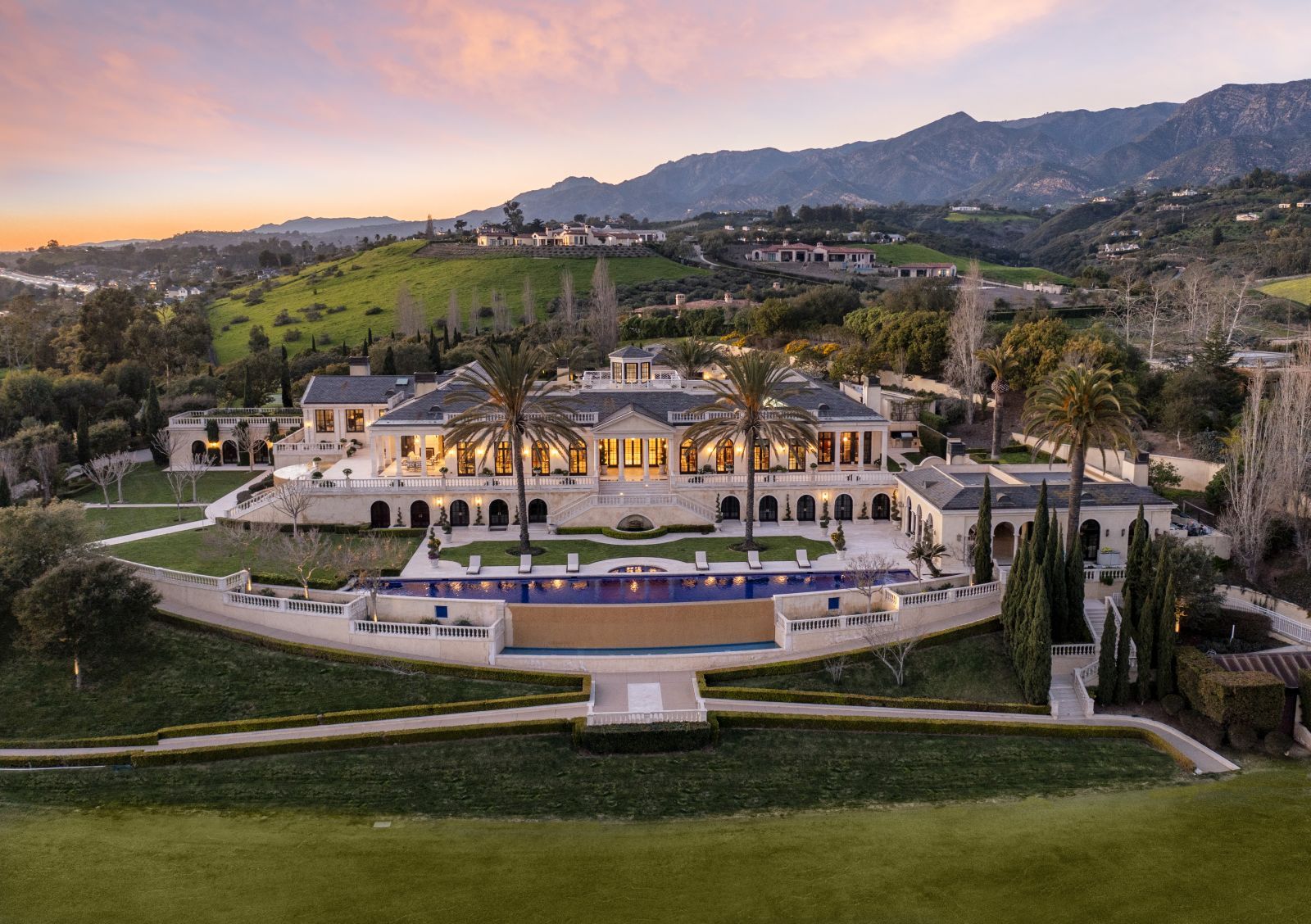 Birds eye view of a 28,000 sq.ft. estate home, with palm tress, swimming pool and expansive green lawn in the foreground and mountains at sunset in the background.