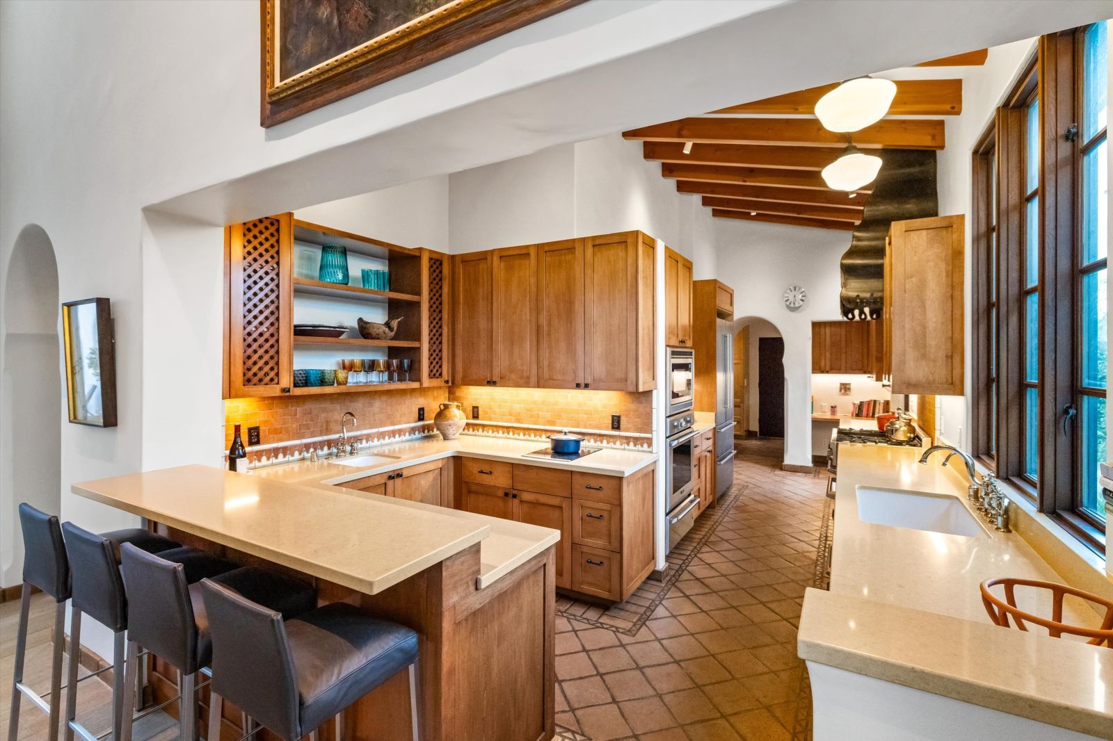 A galley-style kitchen with terracotta tile flooring; high, open wood beam ceiling; and wood-stained cabinets accented with black hardware.