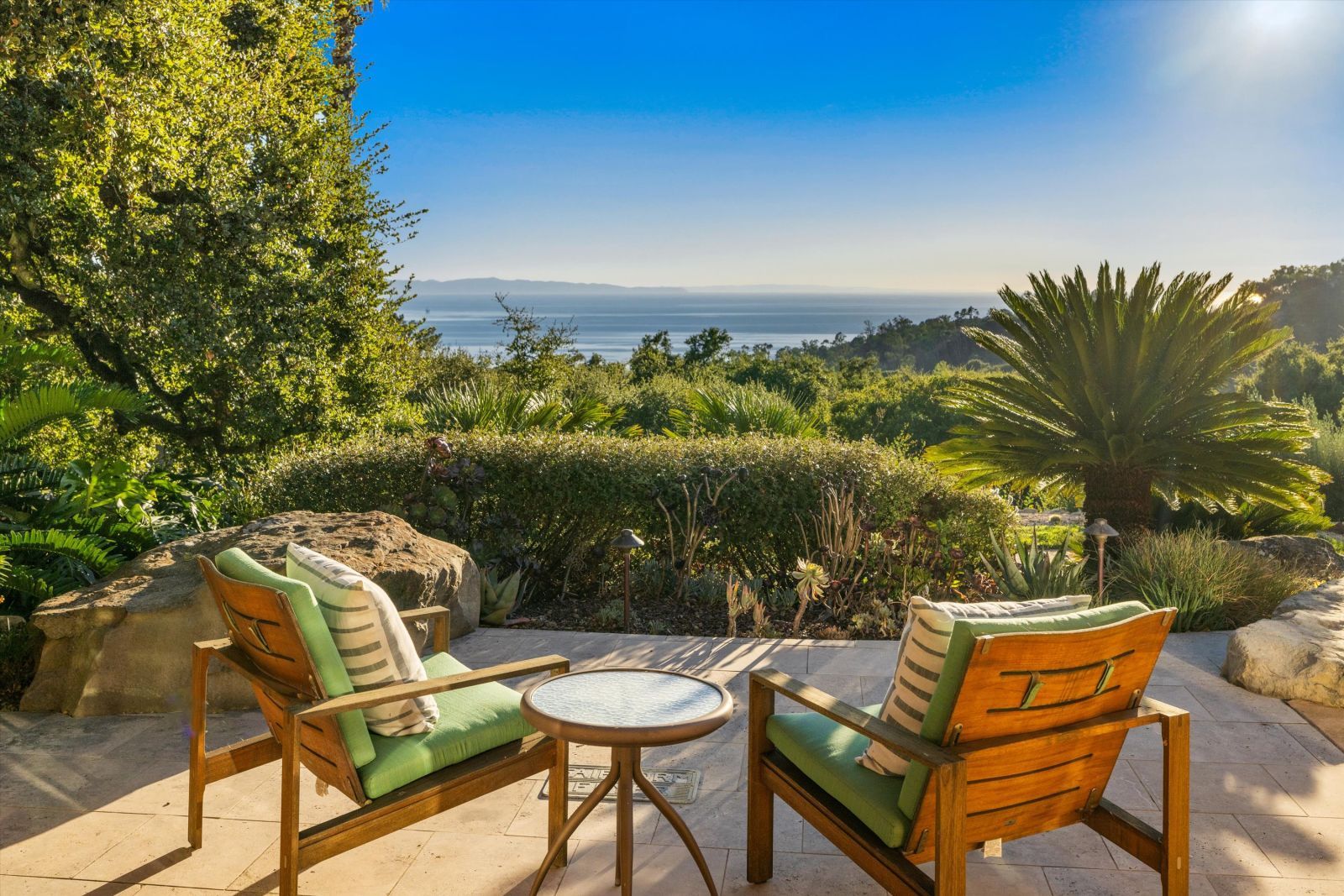Two deck chairs and small round table on a patio overlooking lush greenery and the sparkling ocean on a sunny day.