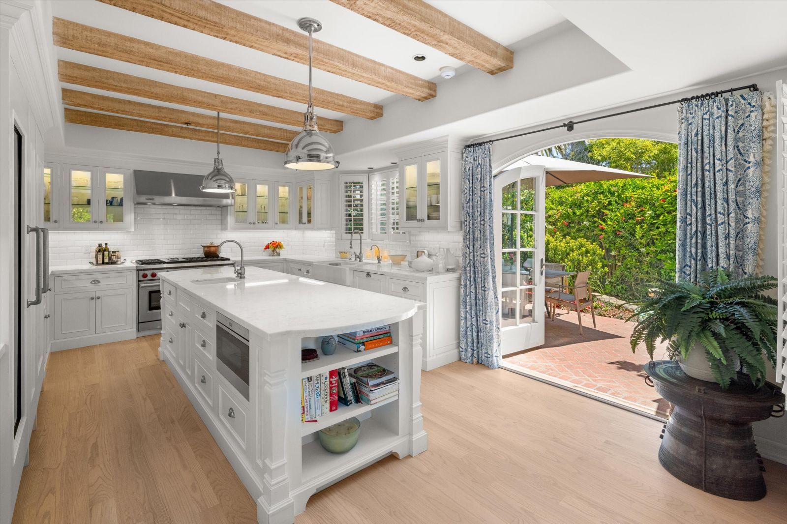 A sunny white kitchen with wood floor, wood ceiling beams, and a center island, with open French Doors leading to the backyard.