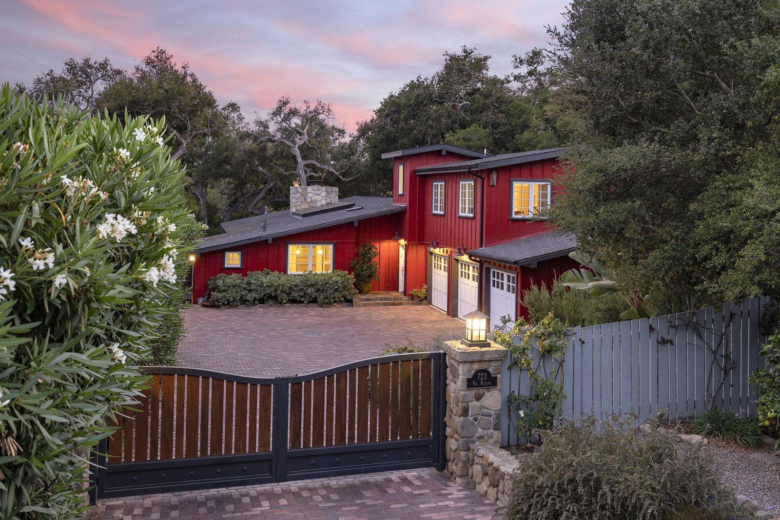 A wooden gate leads to a red farmhouse surrounded by lush foliage, with a pink hued sunset in the background