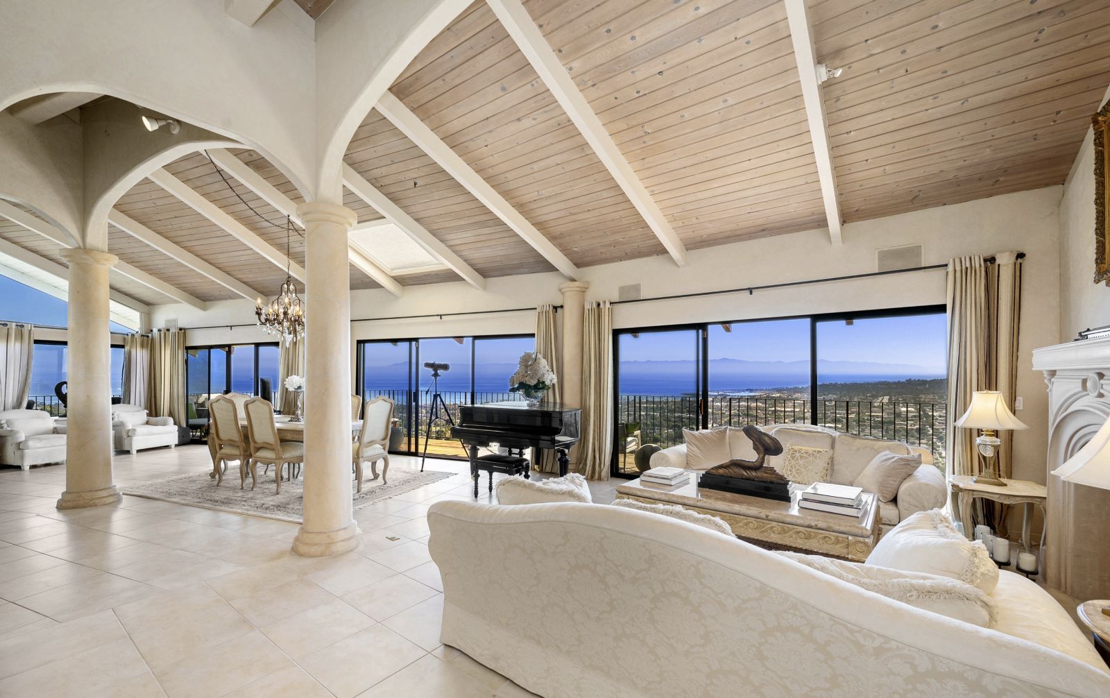 The all-white living room of a Santa Barbara home with columns, beamed ceiling, which plush furniture and windowsa wall of windows looking out to the ocean.