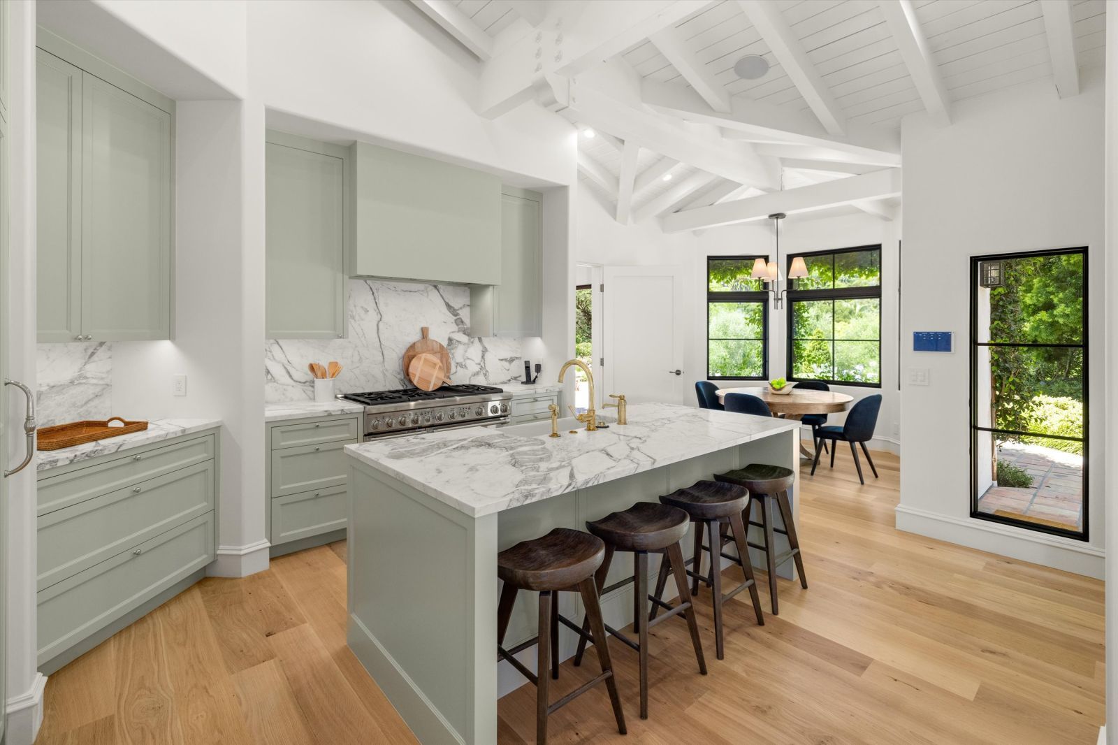 A white kitchen with sage green cabinets, marble countertops, and island stools, with a breakfast room in the background