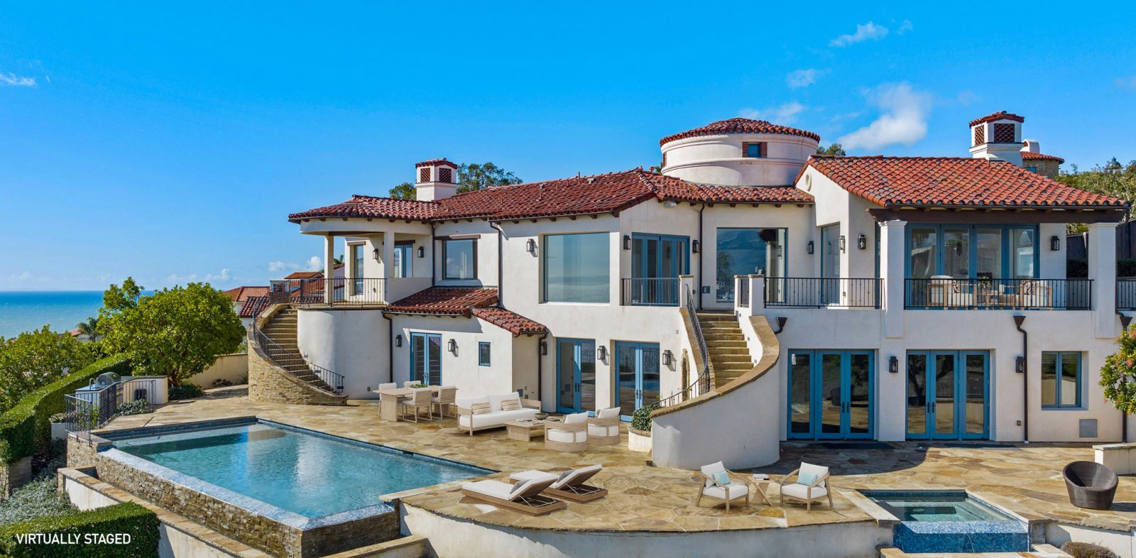 A large, two-story Mediterranean home with white stucco walls, a red tile roof, terraces, and a sparkling blue pool.