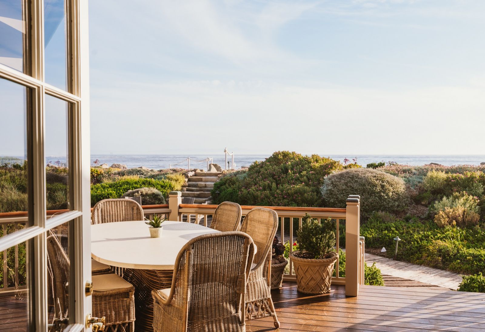 Wicker chairs surround an outdoor table on a beach house terrace overlooking foliage with the beach in the background