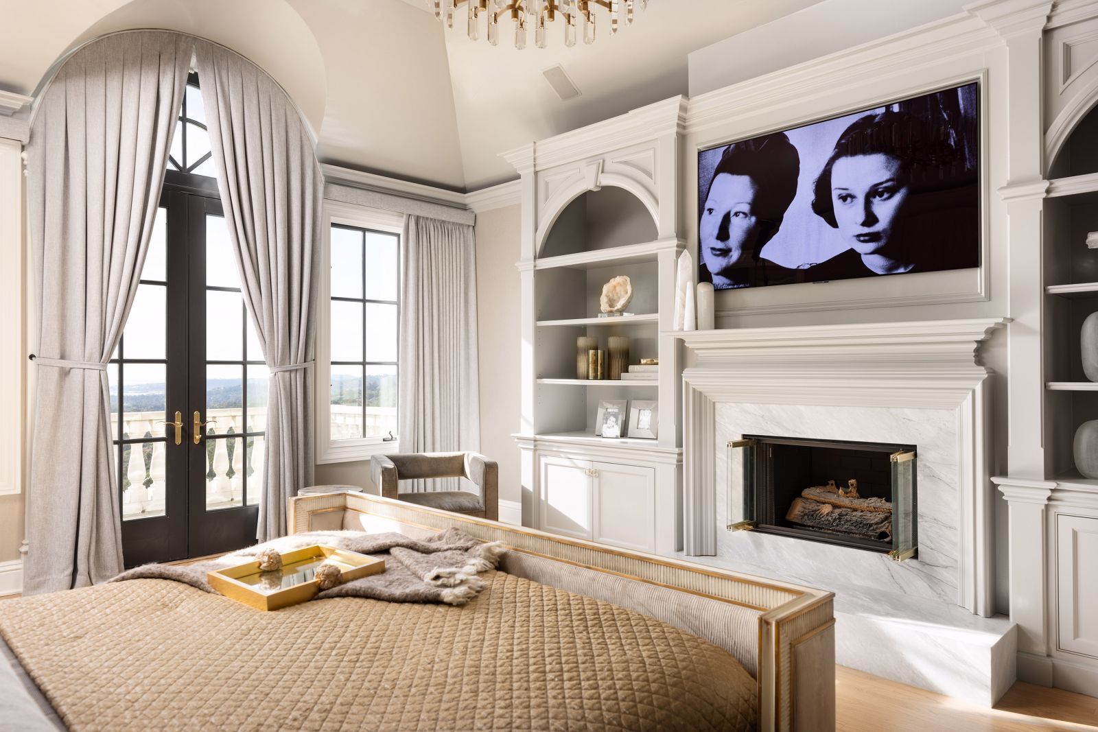 Primary bedroom of a luxurious Montecito home, with a tv above the fireplace, expansive windows looking out to a terrace and the ocean, and French doors