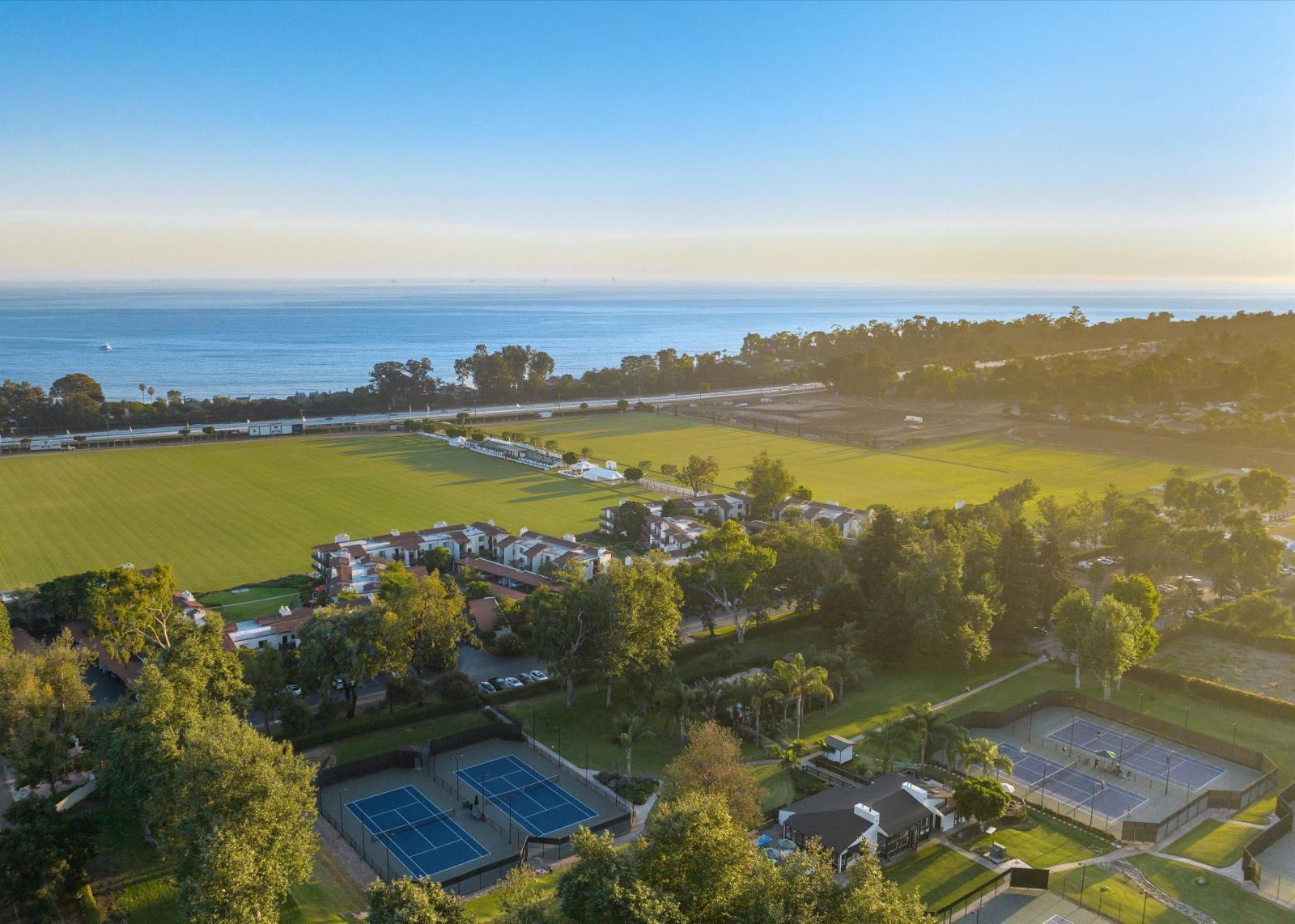 Aerial view of pickelball and tennis courts at the Santa Barbara Polo & Racquet Club, surrounded by lush greenery with the ocean in the background.