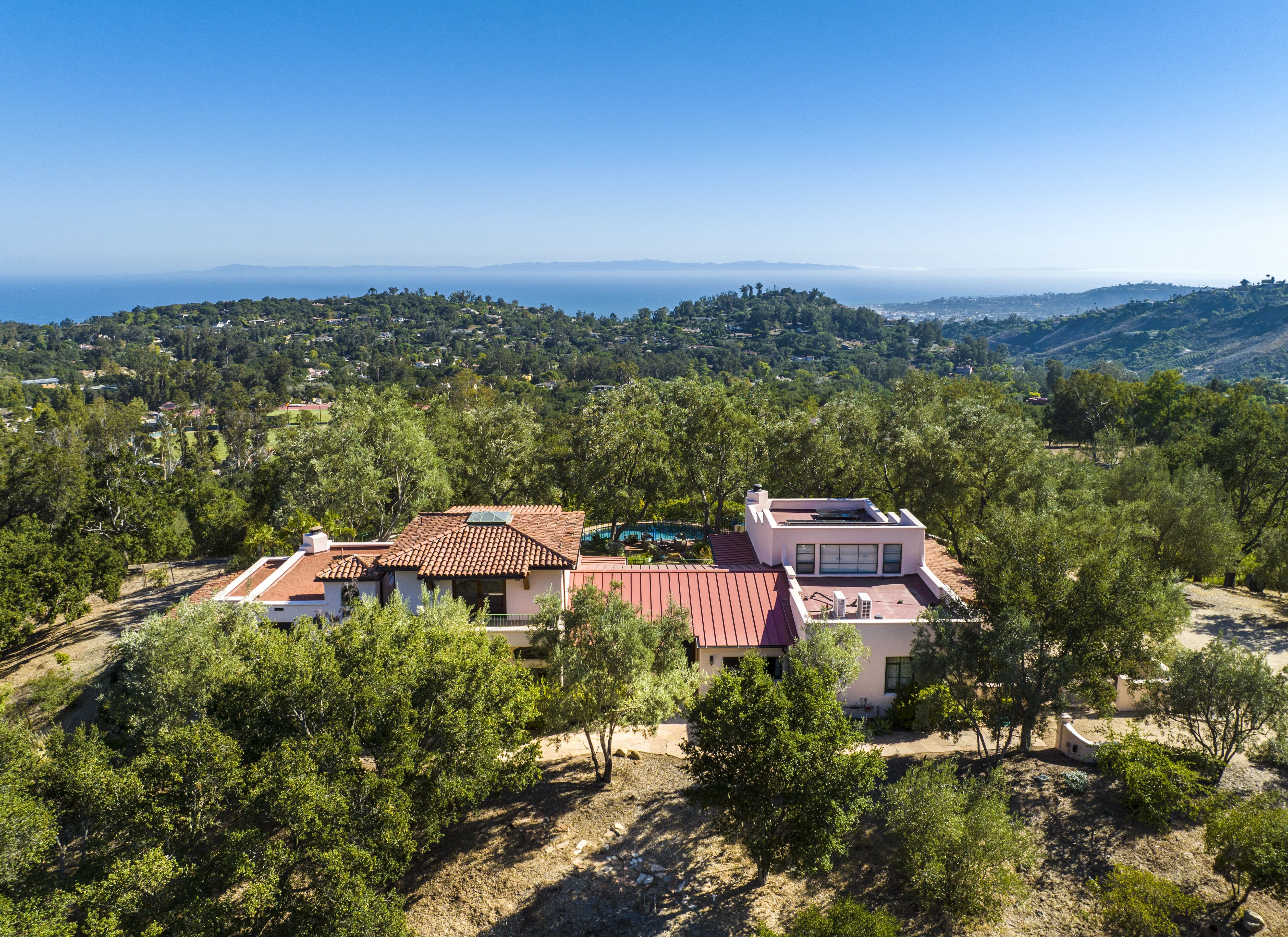 Birds eye view of a Santa Barbara hacienda, with red tile roof, and surrounded by trees, and with the ocean in the distance