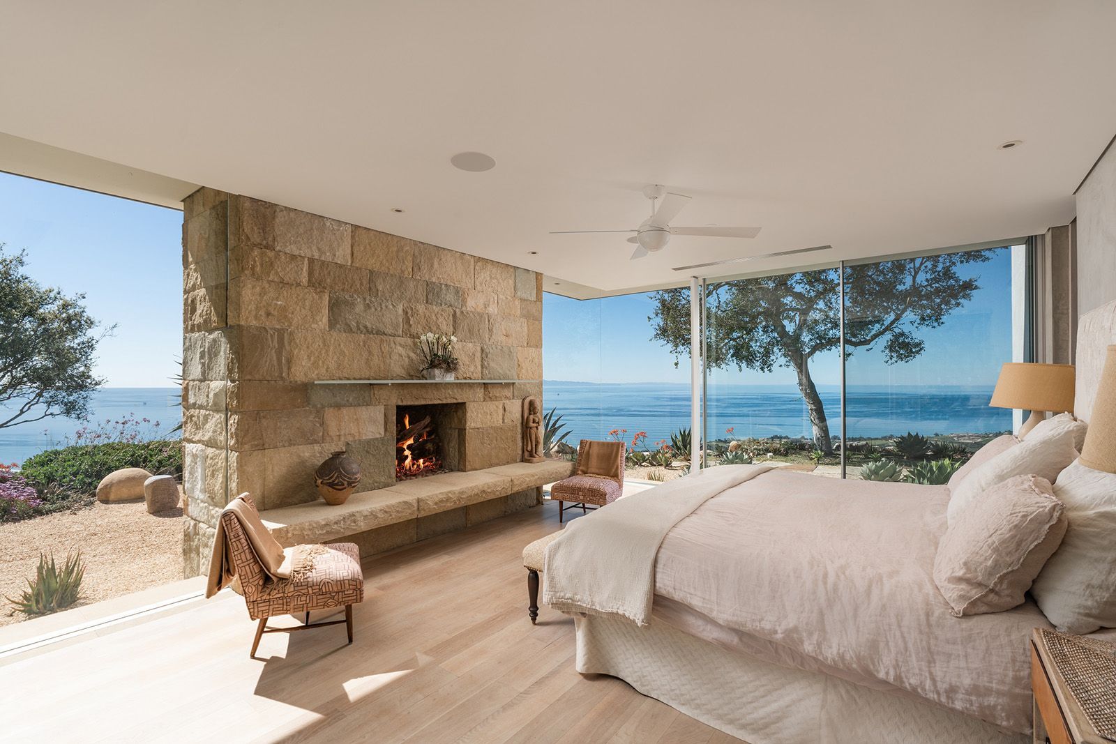 A luxury bedroom surrounded with floor-to-ceiling stone fireplace flanked by walls of windows looking out to the ocean and horizon.
