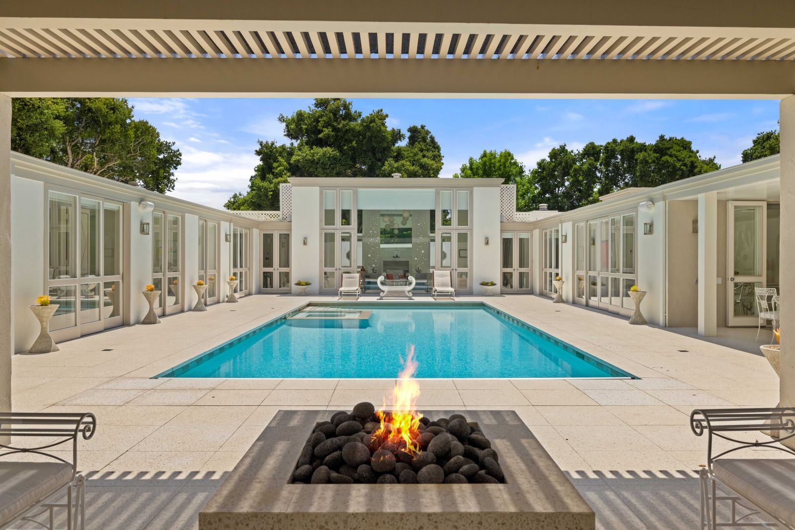 A captivating pool court inside a mid-century home, with a lapiz blue pool in the center, surrounded by deck and glass-fronted rooms of the home. A fire pit is ablaze in the foreground.