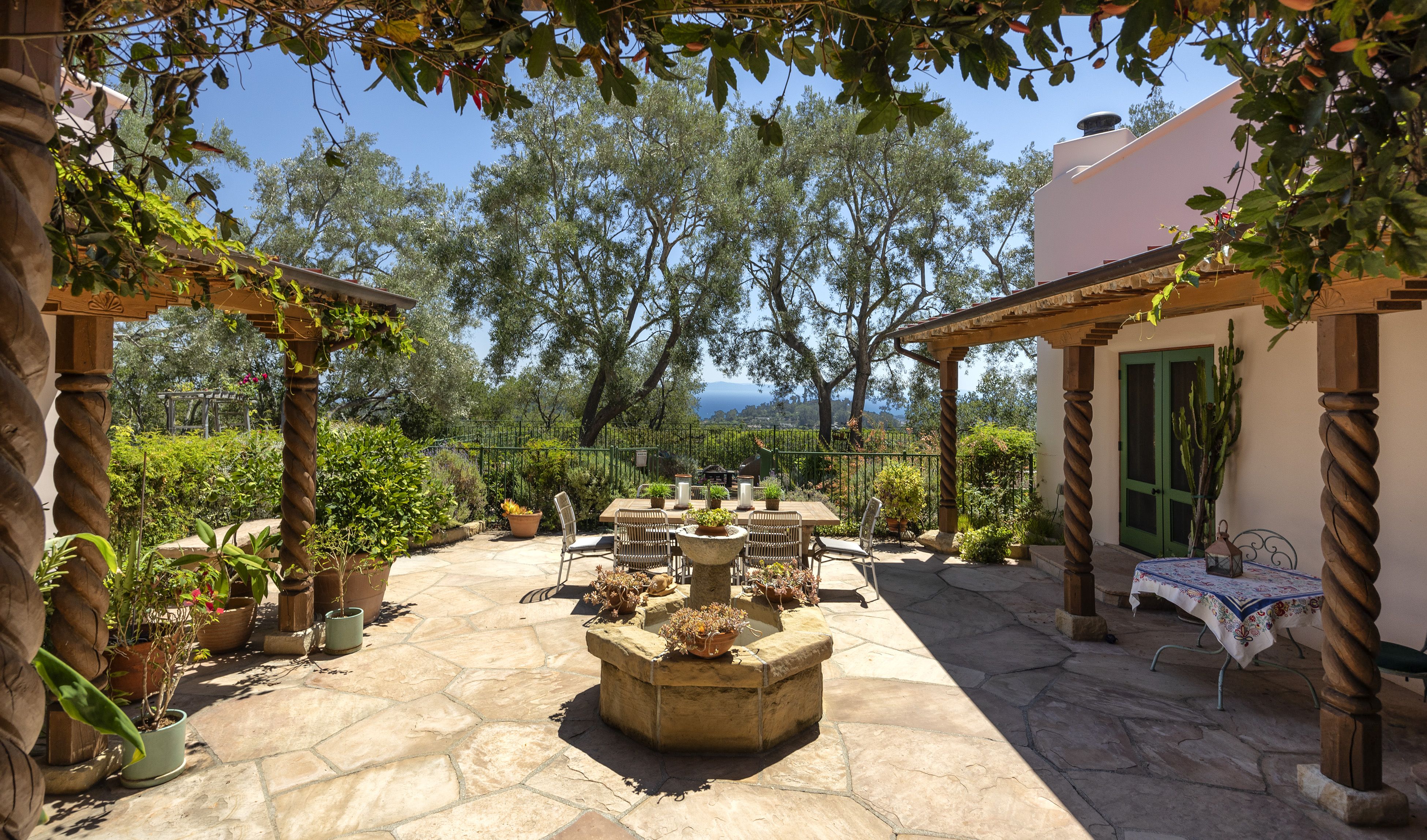 Stone-paved courtyard with fountain, flanked by vine-covered pergolas, with trees and an ocean view in the background