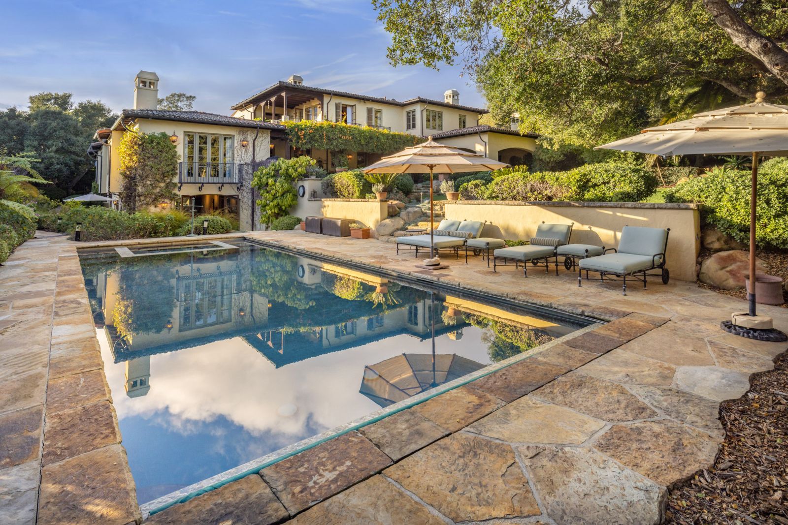 The inviting backyard of a Mediterranean luxury home with a sparkling swimming pool surrounded by patio furniture and umbrellas.