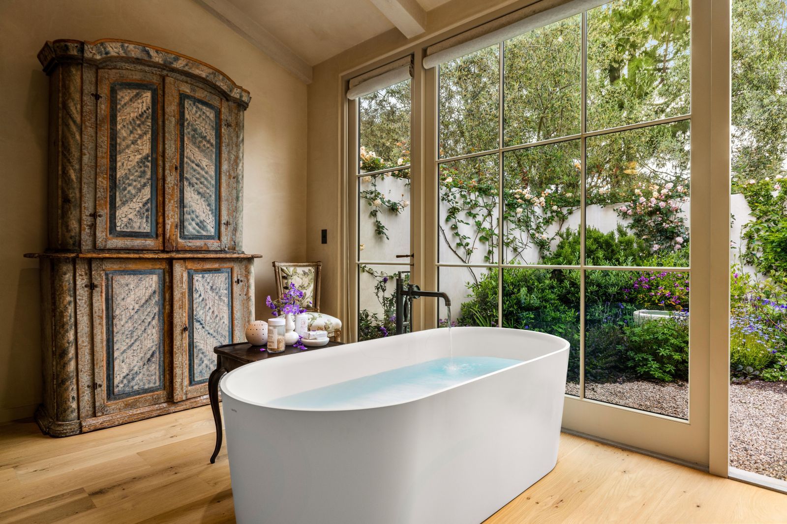 A bathroom with a free standing soaking tub in front of a multi-pane wall-size with lush vegetation and an armoire in the background.