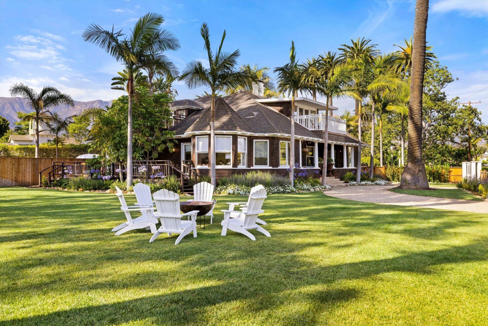 A 1920s era Craftsman style Montecito beach home with expansive lawn, multiple palm trees, and a set of white Adirondack chairs in the foreground