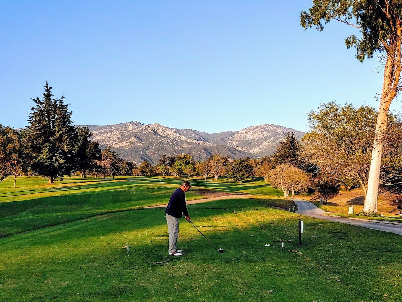 A man in a black sweater sets up to tee off on a hole at Santa Barbara Golf Club, with mountains and blue sky in the background