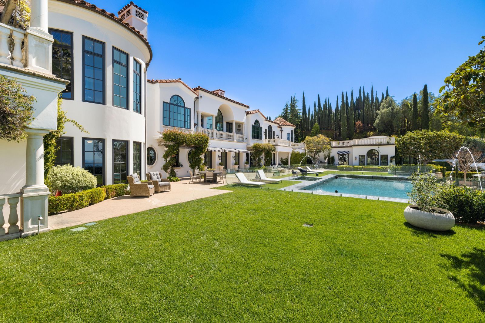 The backyard of a spectacular Montecito estate with green grass surrounding a pool, with a portion of the Meditshowingerranean style home