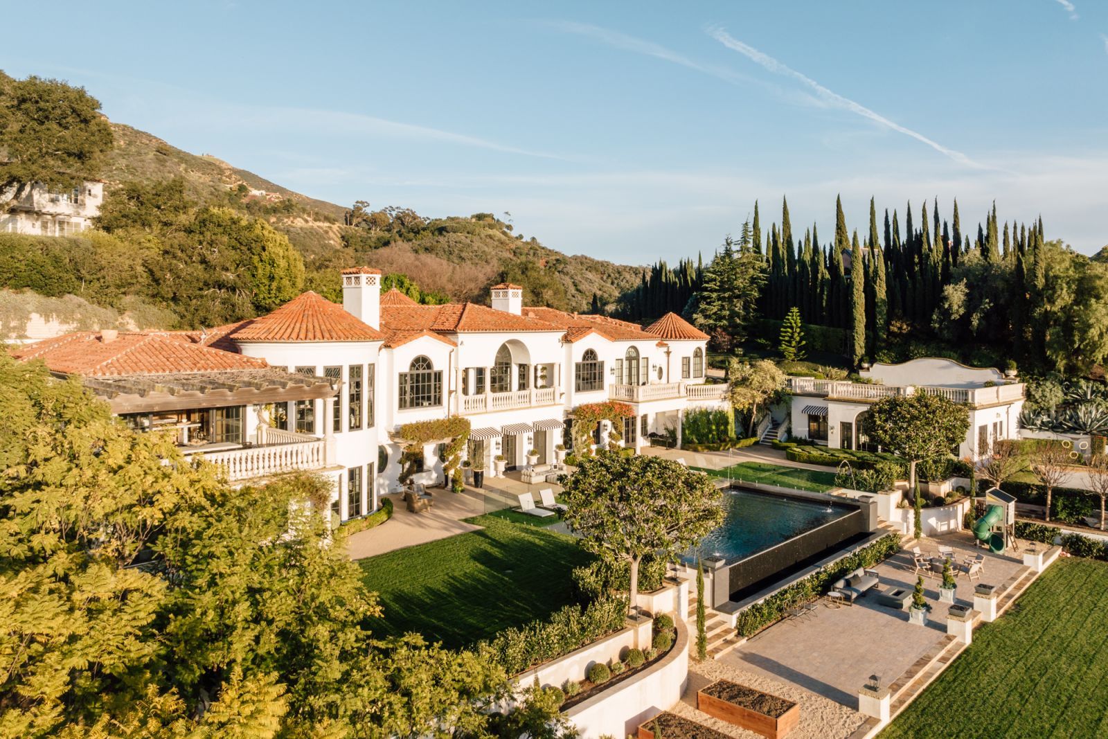 A large multi-level estate home in Montecito with stark white walls and red tile roof, with its beautifully manicured grounds in the foreground.