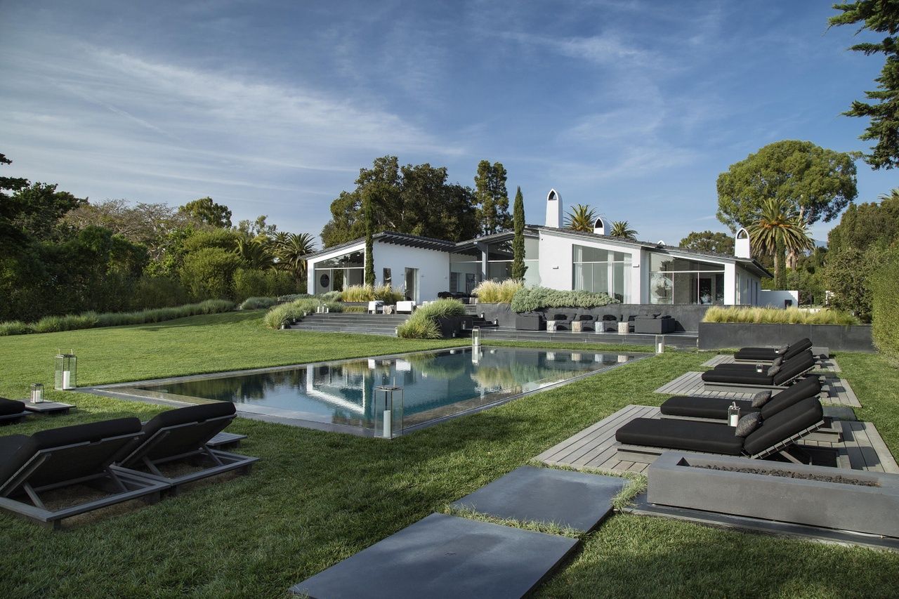 The rear grounds of a Hope Ranch estate with a swimming pool, lots of grass, lounge chairs and the white stucco home in the background under a blue sky.