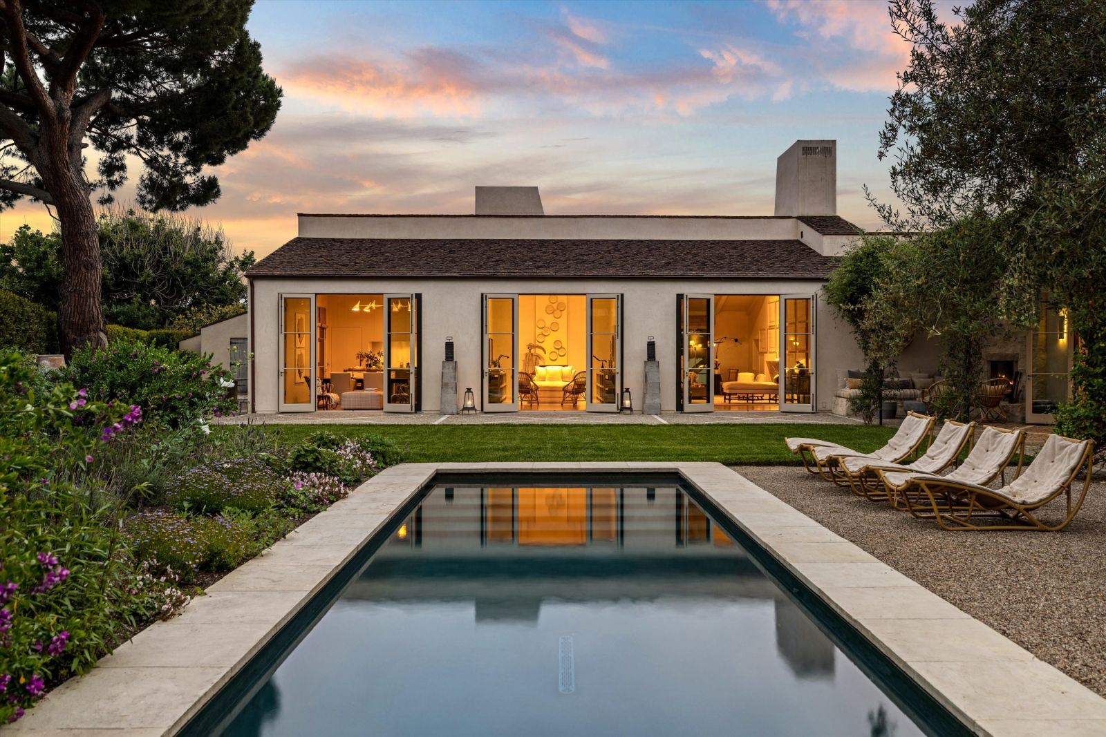 Backyard of a home at sunset, with floor to ceiling windows illuminated from within, with a pool, lounge chairs and lawn in the foreground.