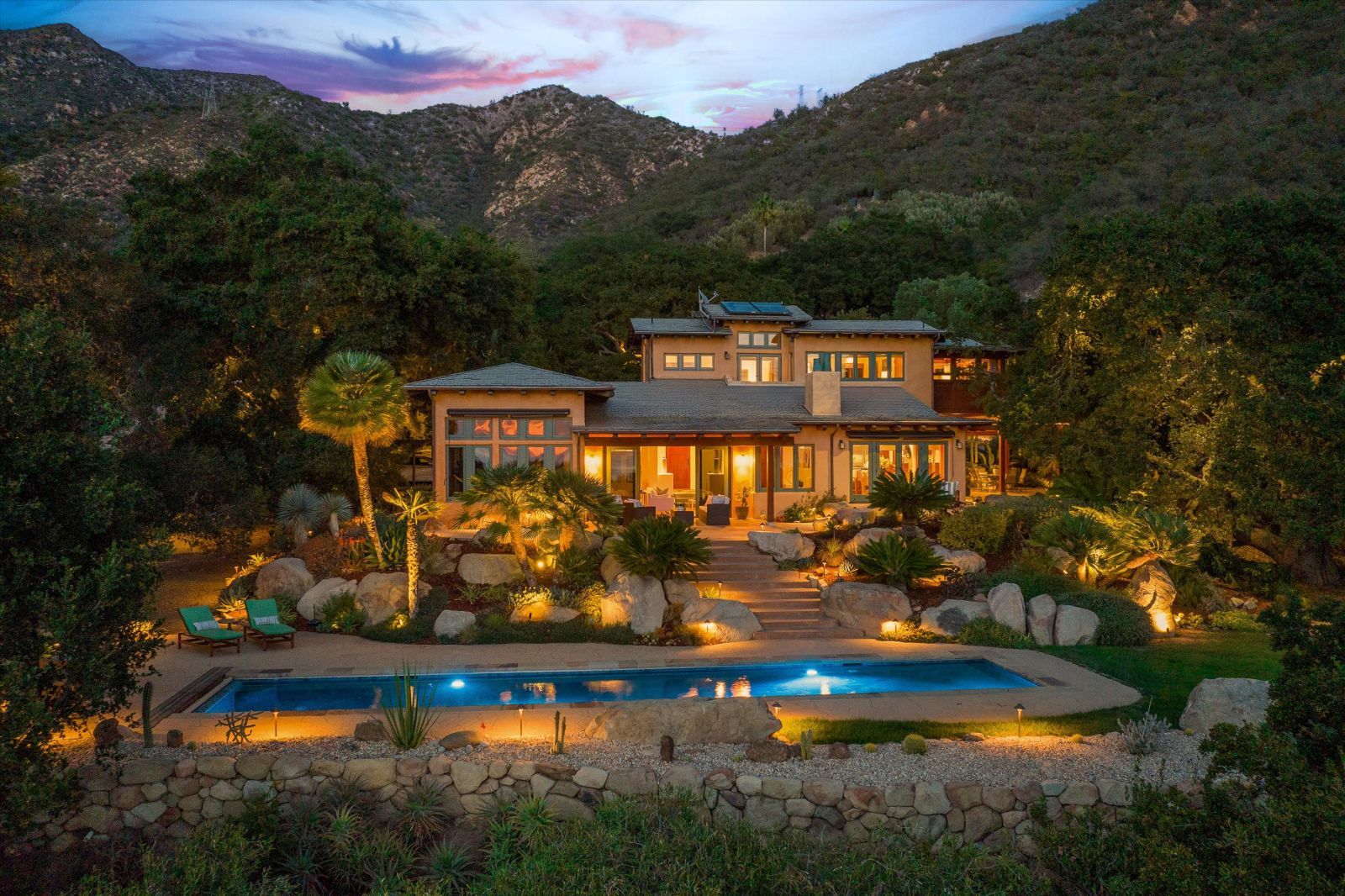 A Craftsman inspired, Mediterranean influenced home at dusk, set against a hillside with mountains in the background,illuminated from within, and with a beautiful backyard with lighted pool.