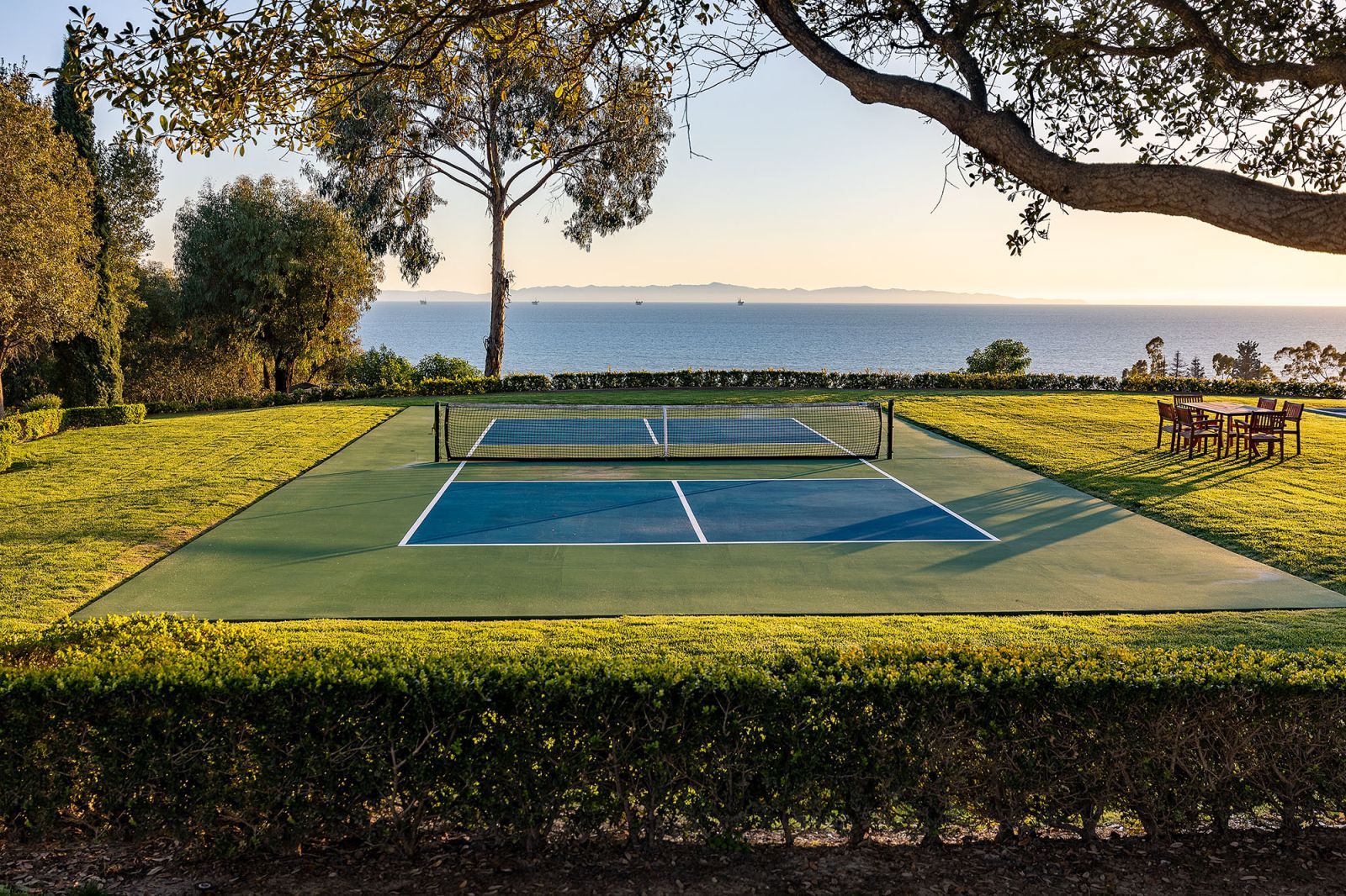 The verdant grounds of an estate, looking toward a pickleball court surrounded by verdant lawn, with the blue ocean in the background