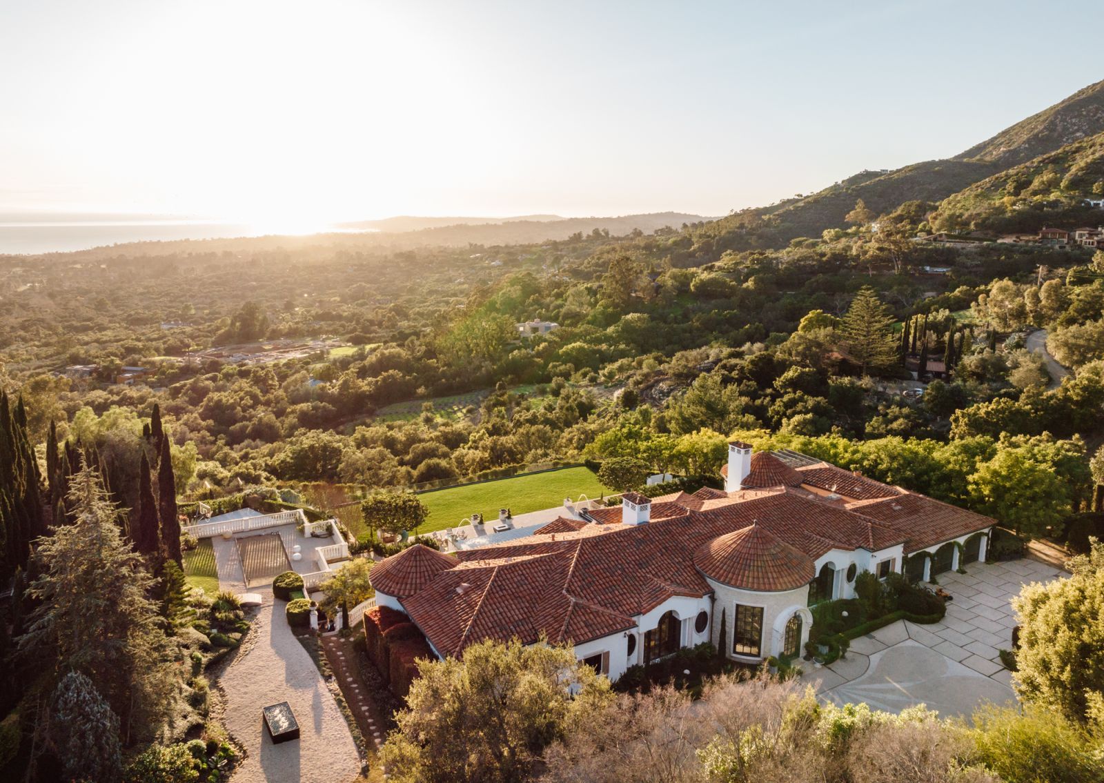 A bird's eye view of a trophy home in Montecito featuring the red tiled roof, surrounding lush foliage, and the ocean on the horizon