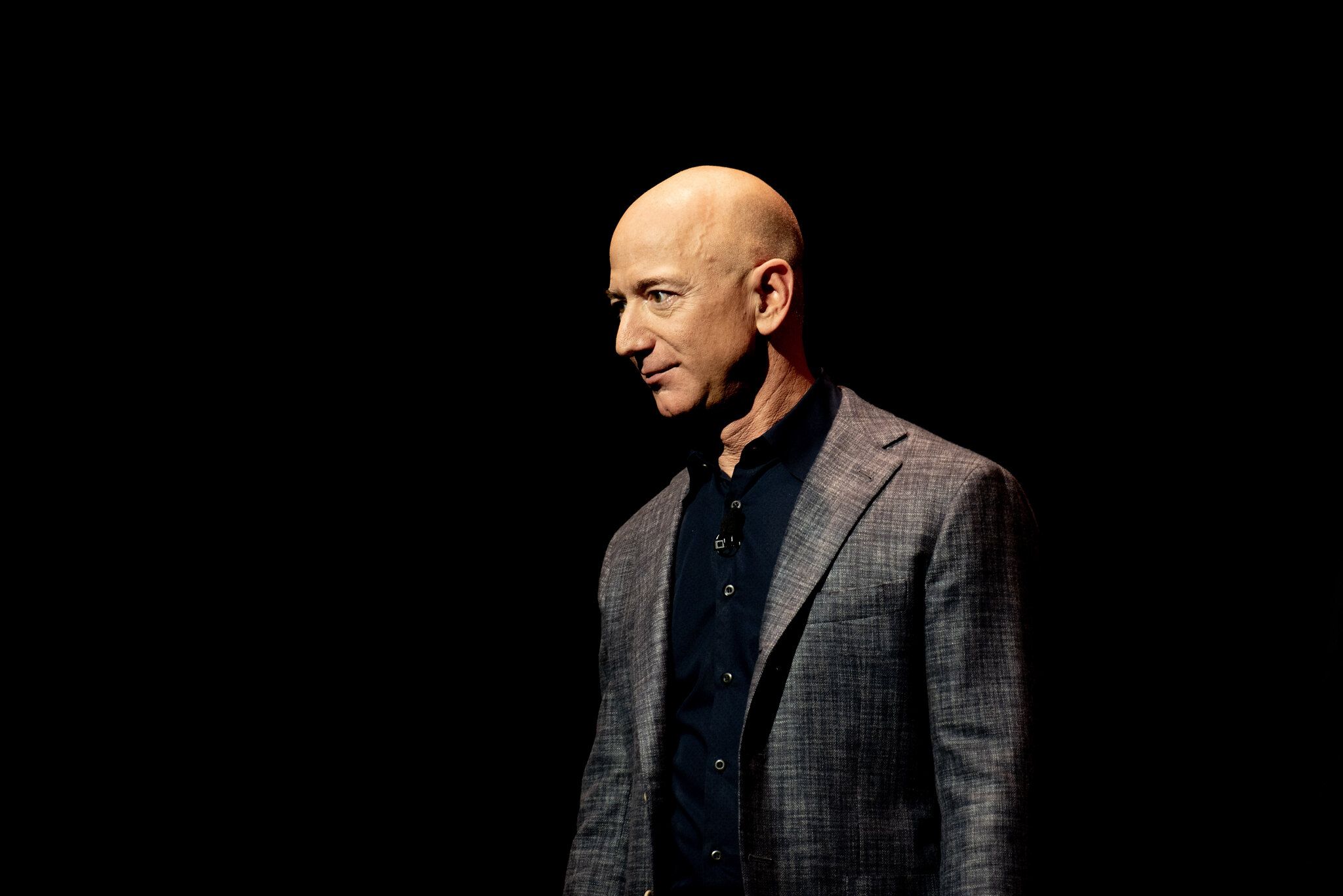 Jeff Bezos in a suit and open-neck black shirt in front of a black background.