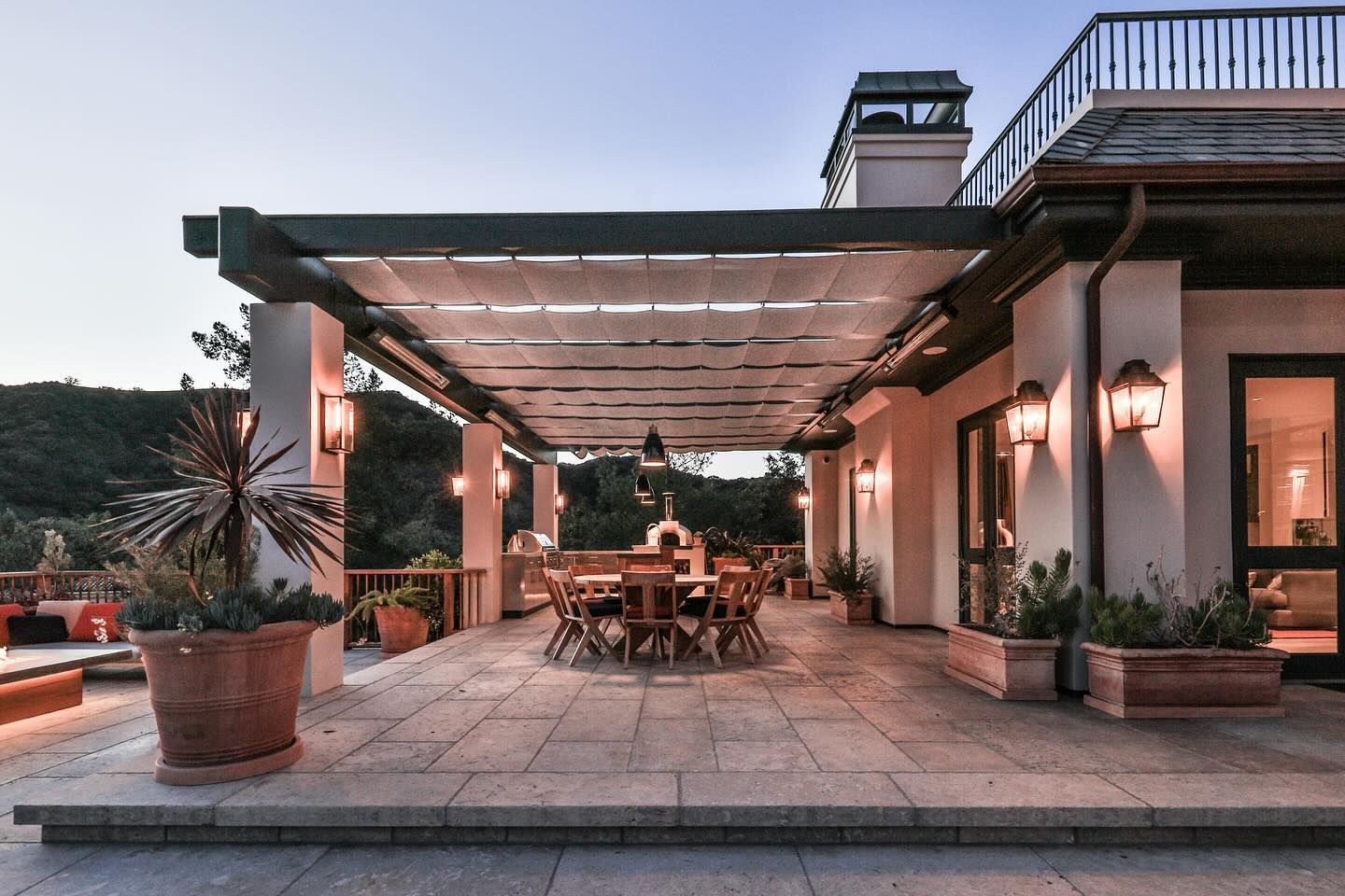 Expansive stone terrace covered by a pergola with the glow of the home's interior lighting emanating through windows.