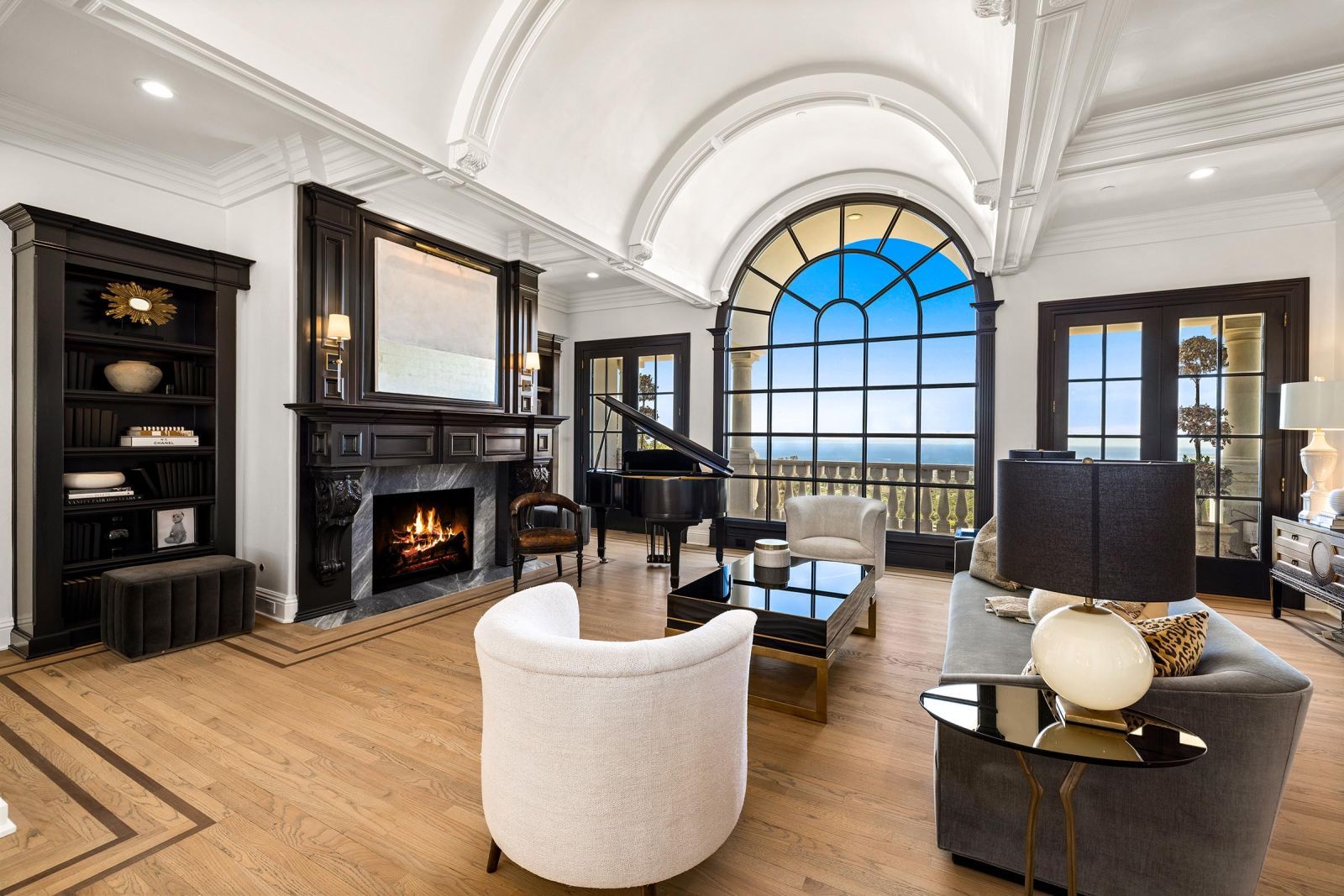 The living room of a luxury home with dark paneling, a huge fireplace, a barrel vaulted ceiling, and a massive multi-paned arched window with an ocean viewy elegant yet comfy furniture