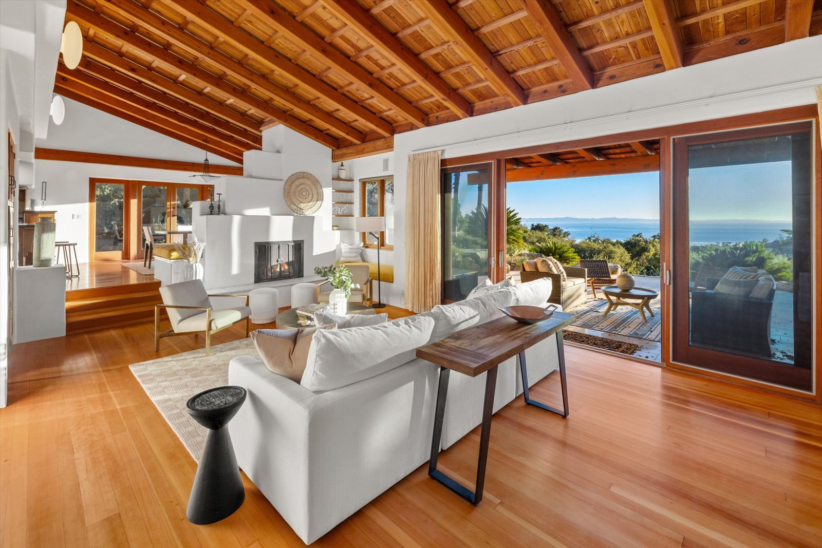 The living room of a luxury home with wood beamed ceiling, wood floor, and white walls, and a breathtaking ocean view.