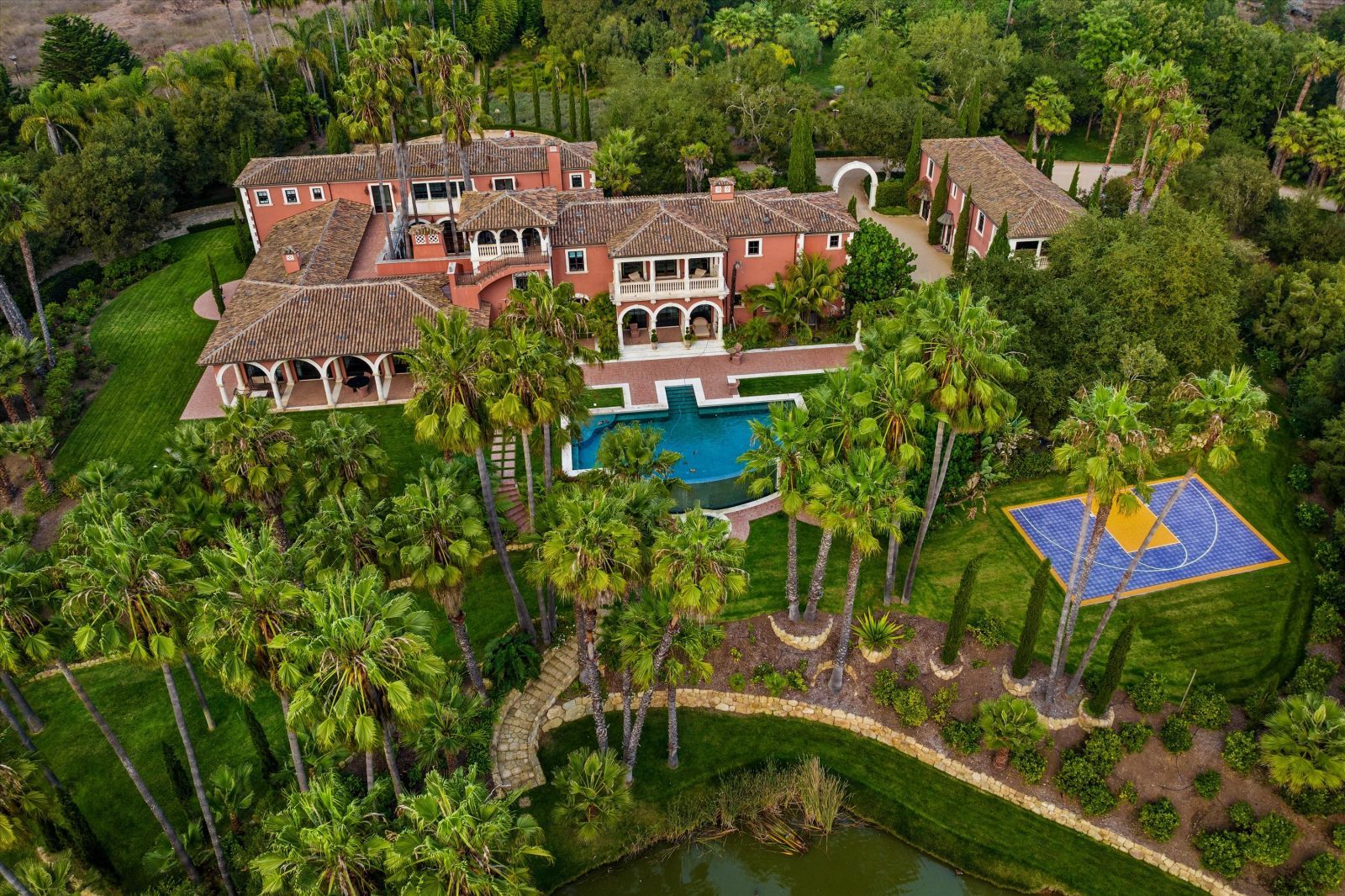 Ariel view of a multi-level luxury Hacienda-style Spanish Colonial home and its swimming pool and tennis court on an estate, surrounded by lush landscaping and mature trees.
