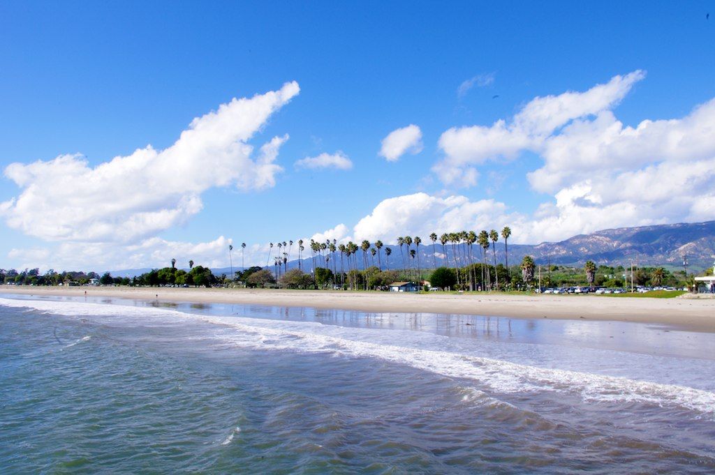 A long stretch of ocean and beach with small waves lapping the shore and palm trees, blue sky and wispy clouds in the distance.
