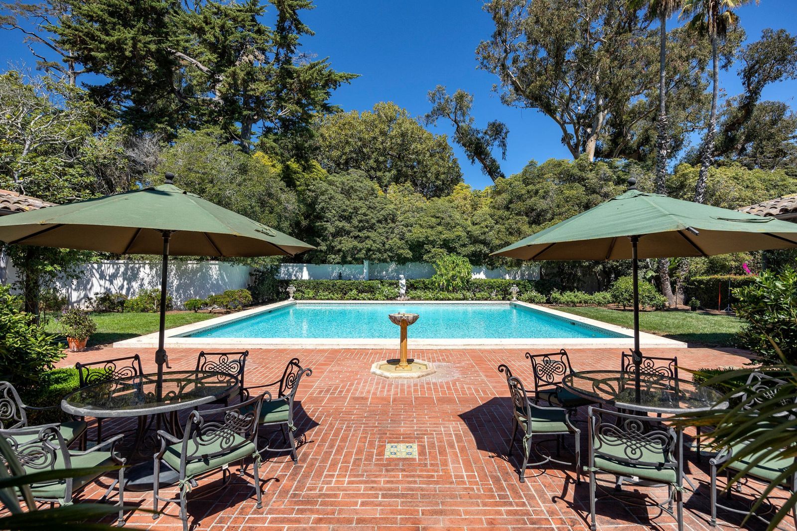 A luxury backyard red brick patio with a serene pool, a small water fountain and two sets of outdoor tables, chairs and umbrellas.