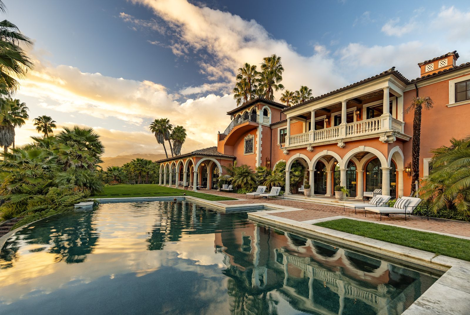 The rear facade of a dramatic ochre-colored Spanish Colonial-style estate home with a sparkling pool under a beautiful sky with fluffy clouds at dusk