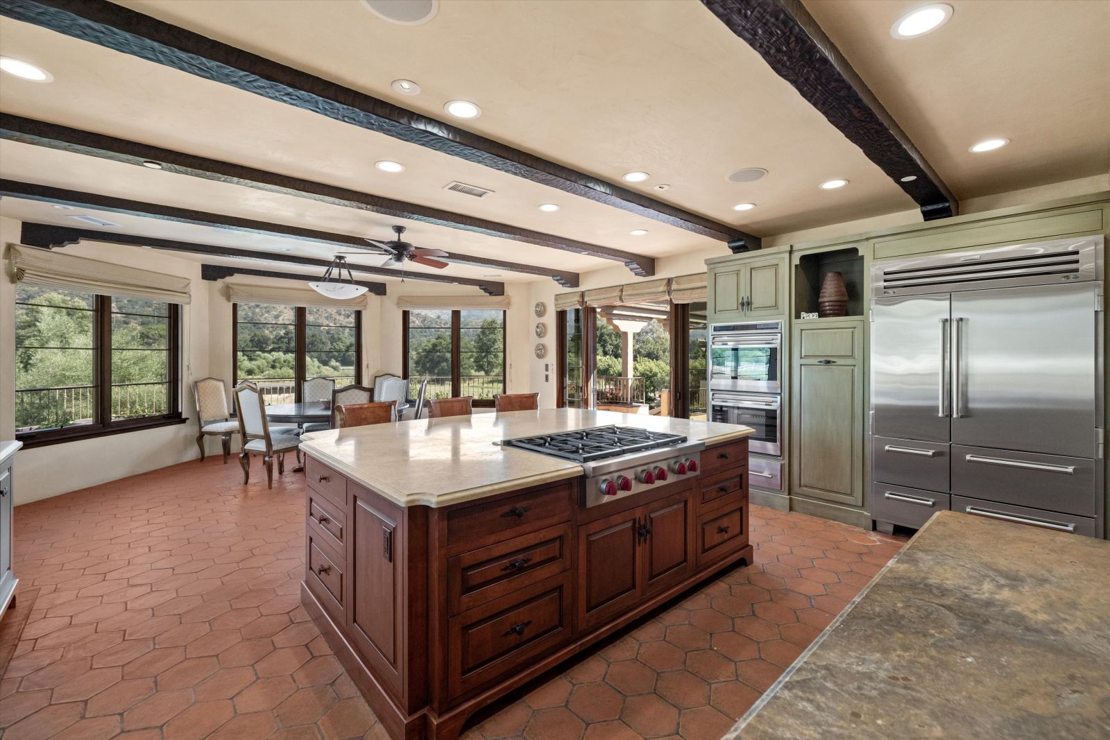 An open concept kitchen an spacious breakfast area, with the kitchen in the foreground hosting a large center island with a stone countertop, stainless appliance, and open wood beam ceiling, and wall of windows behind the dining area.to the room with stainless appliances and a tile floor.
