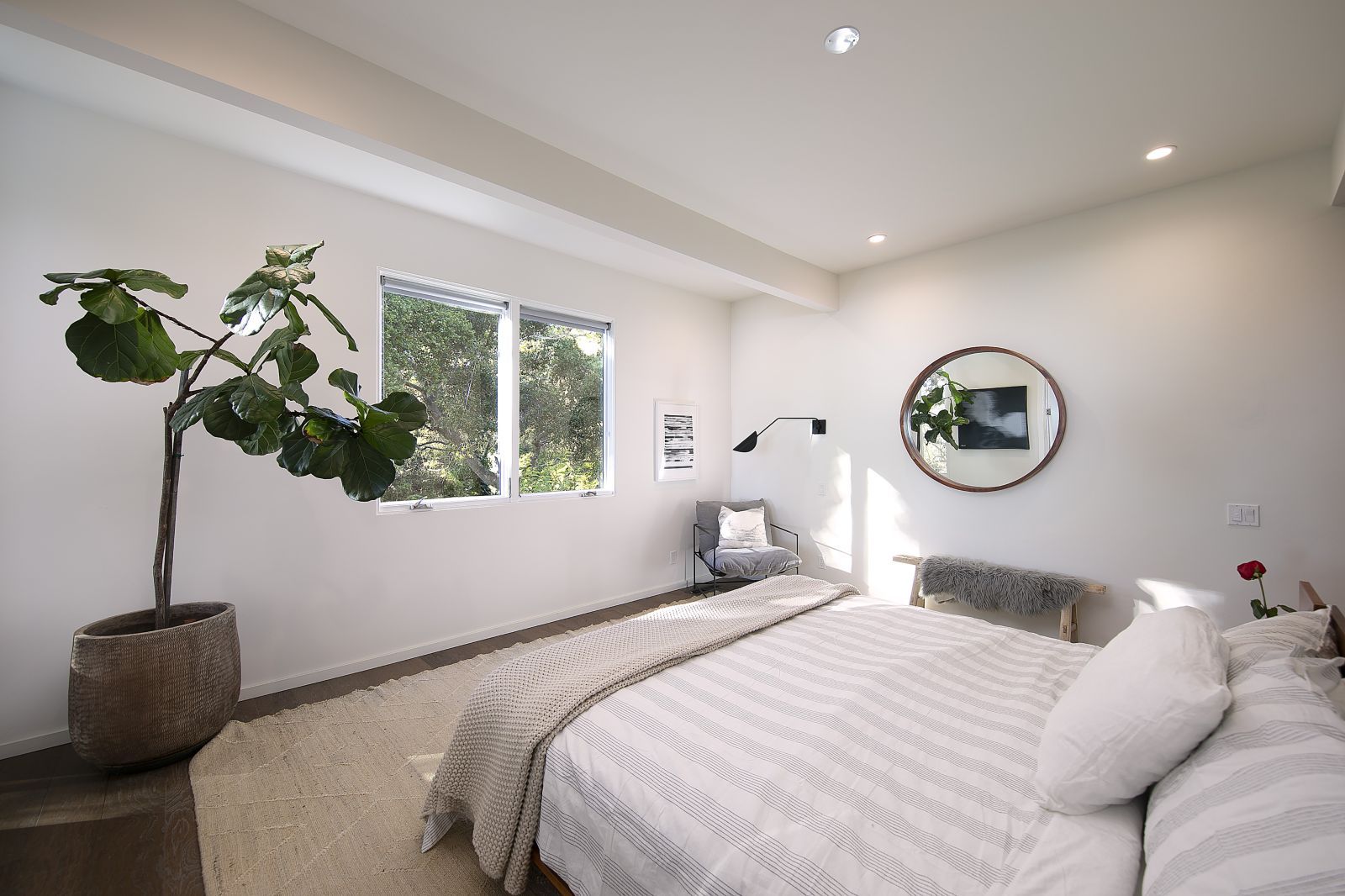 A serene bedroom with a plant in the corner and a mirror on the wall with a modern light fixture above a modern looking chair.