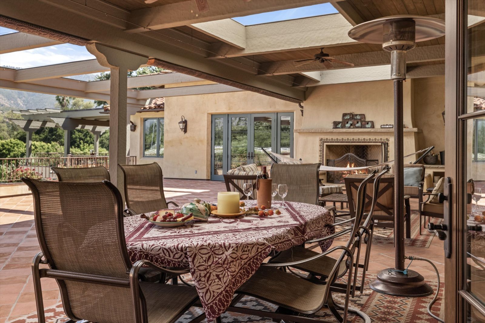 The table is set for al fresco dining on this luxury patio with a lit fireplace, pergola, and hills in the distance.