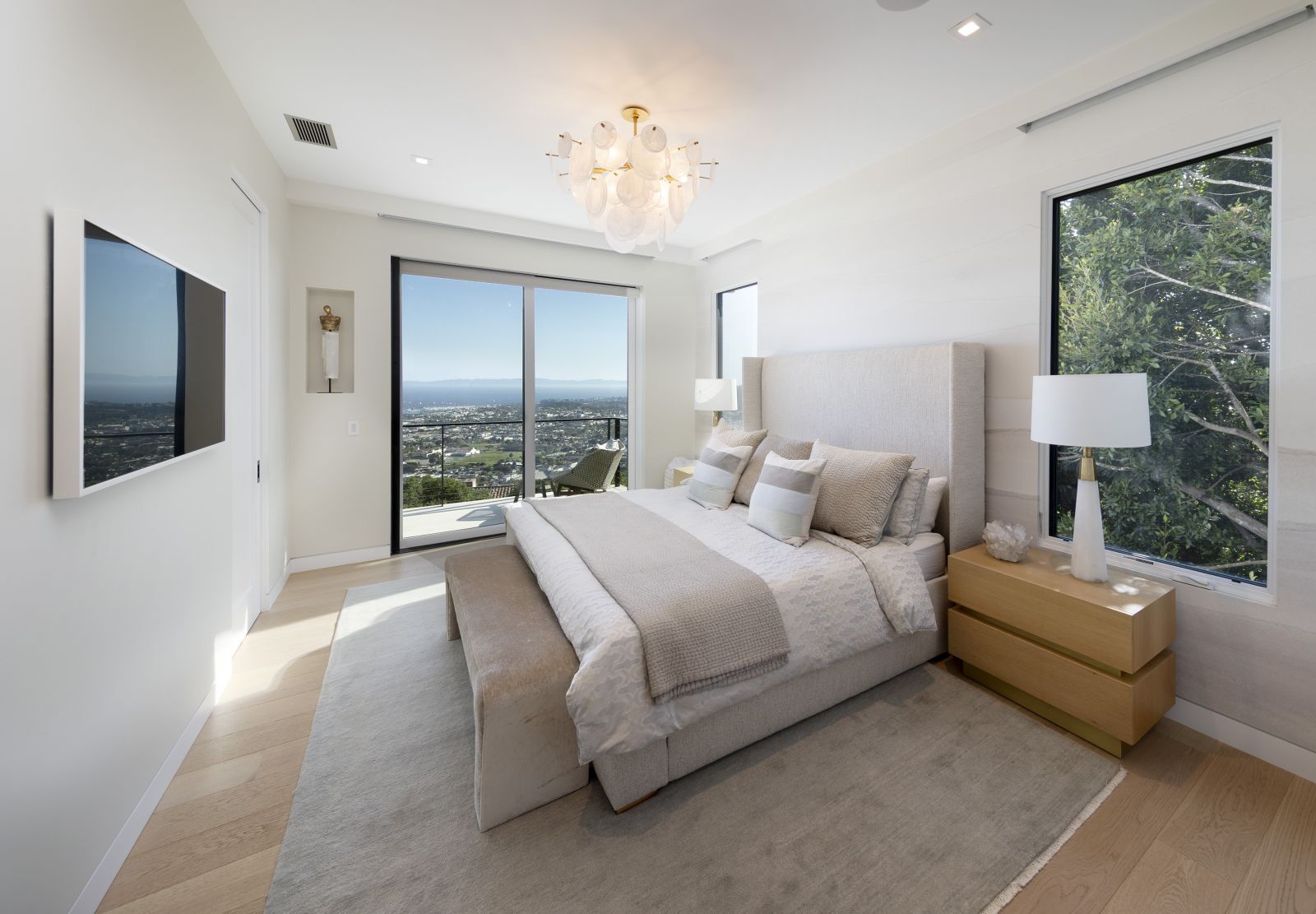 A stylish primary bedroom suite, outfitted in a white and beige color palette, with glass doors that open to city and ocean views.