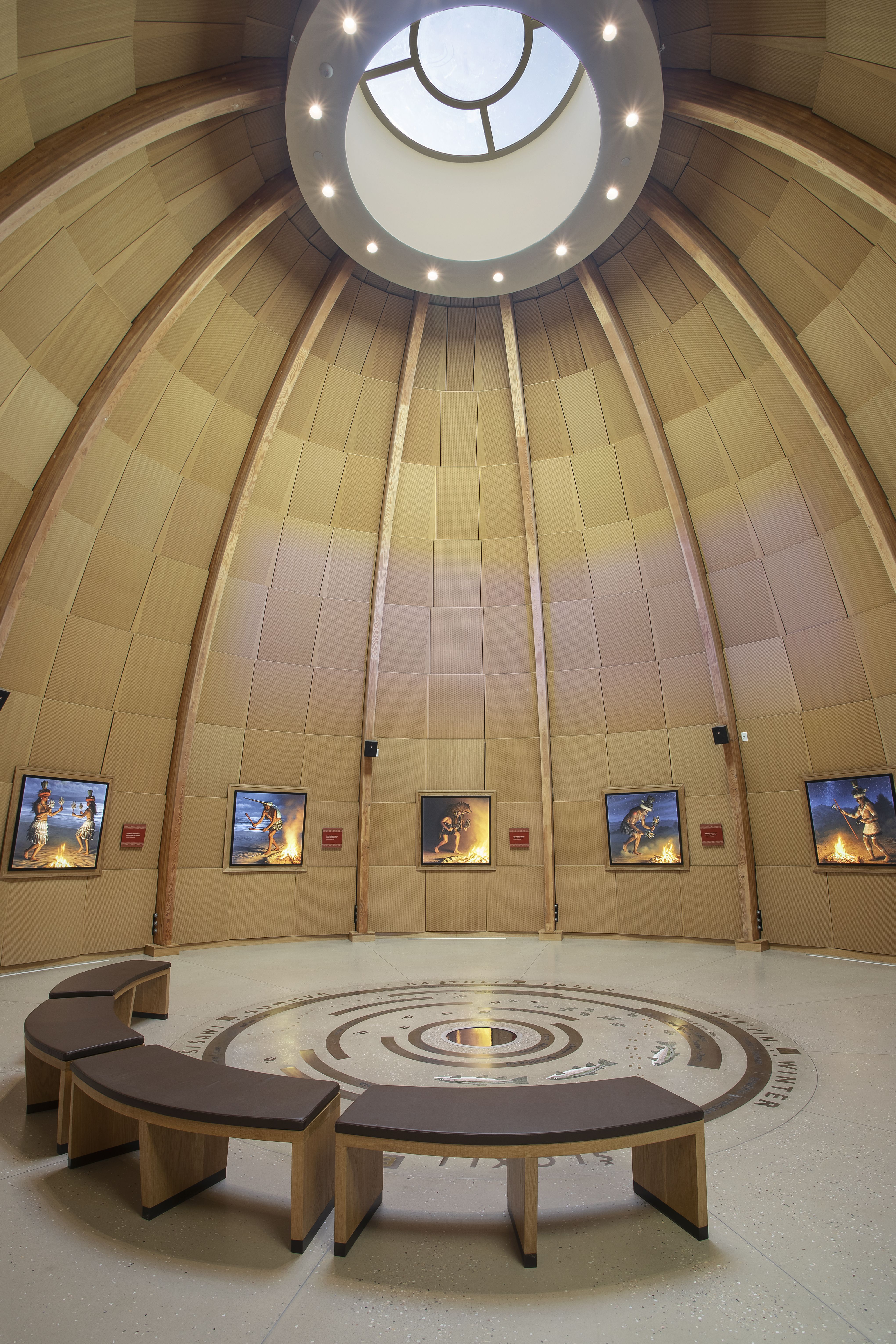 Inside a dome structure with a round skylight in the soaring ceiling, and televisioons along the walls surrounding simple yet elegant benches.