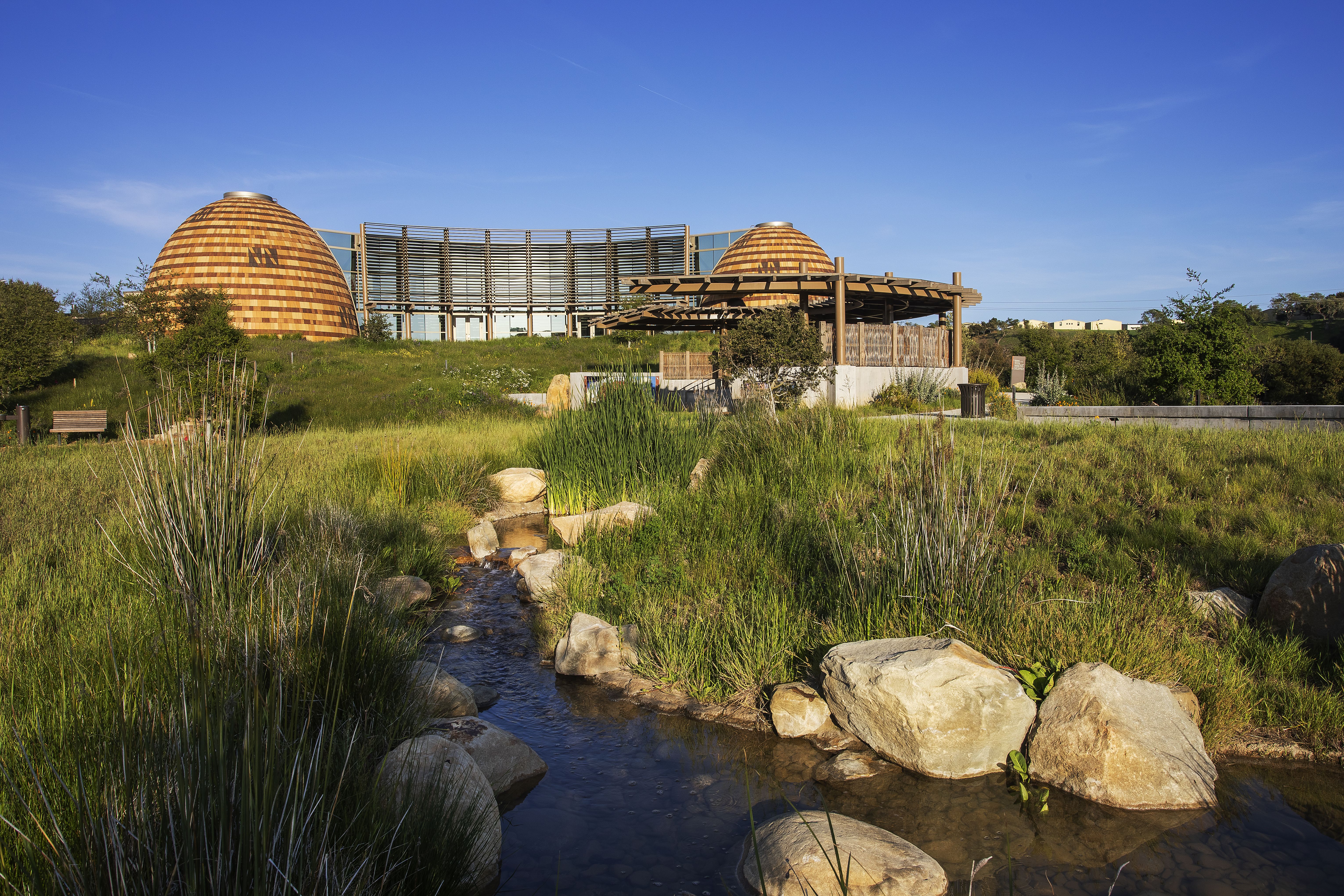 A wide view of the new Santa Ynez Chumash Museum and Cultural Center, with a creek lined with small boulders in the foreground, lush flora, and two Chumash dome structures in the background.