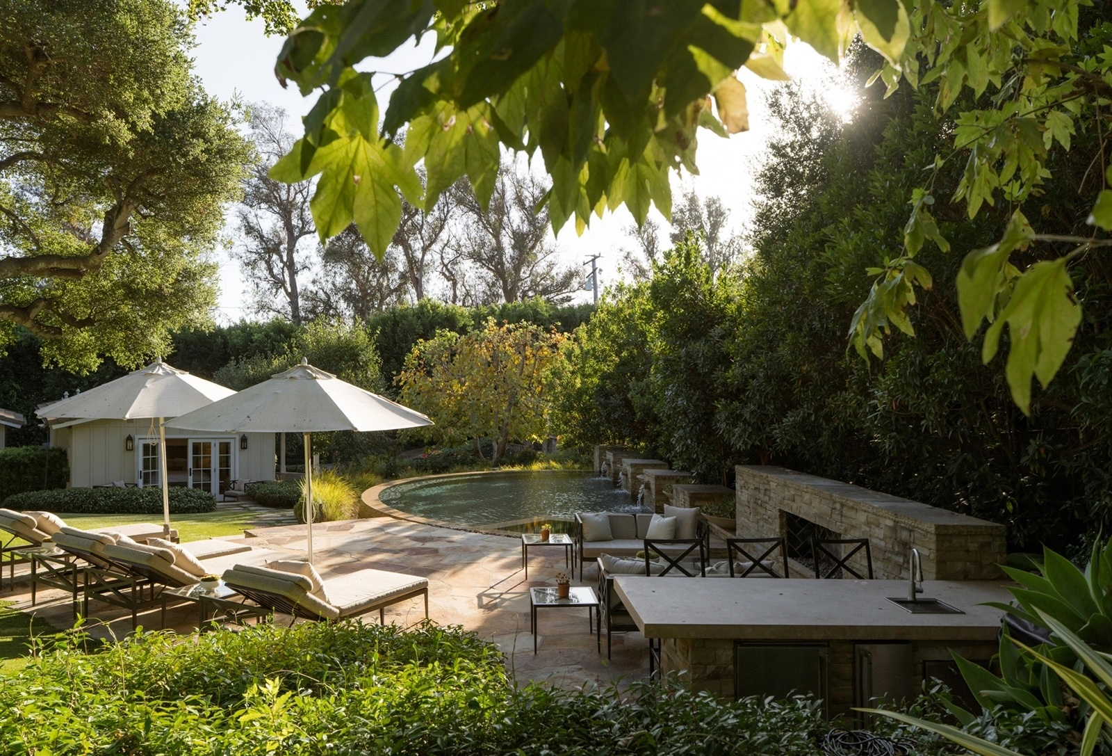 Sunlit backyard retreat with a swimming pool, cascading fountains, outdoor kitchen and fireplace, lounge seating, and lush mature landscaping.