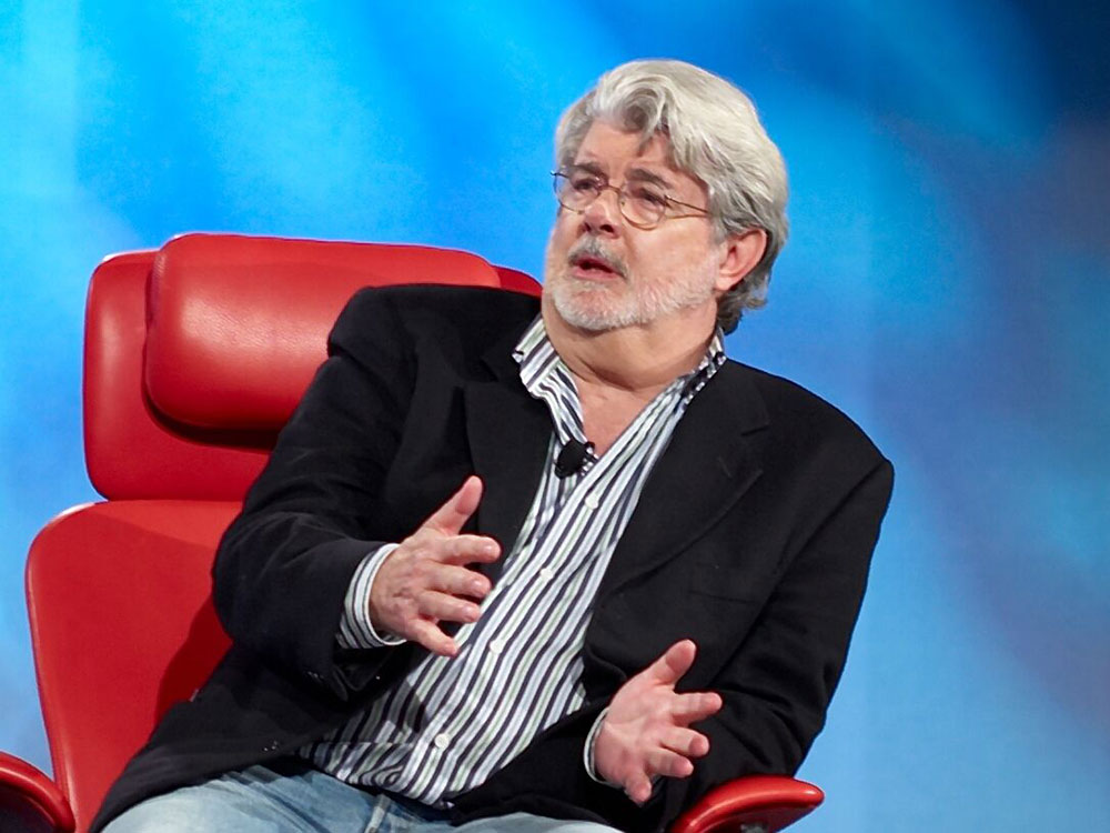 Filmmaker George Lukas sits in a red chair addressing an audience with a blue background