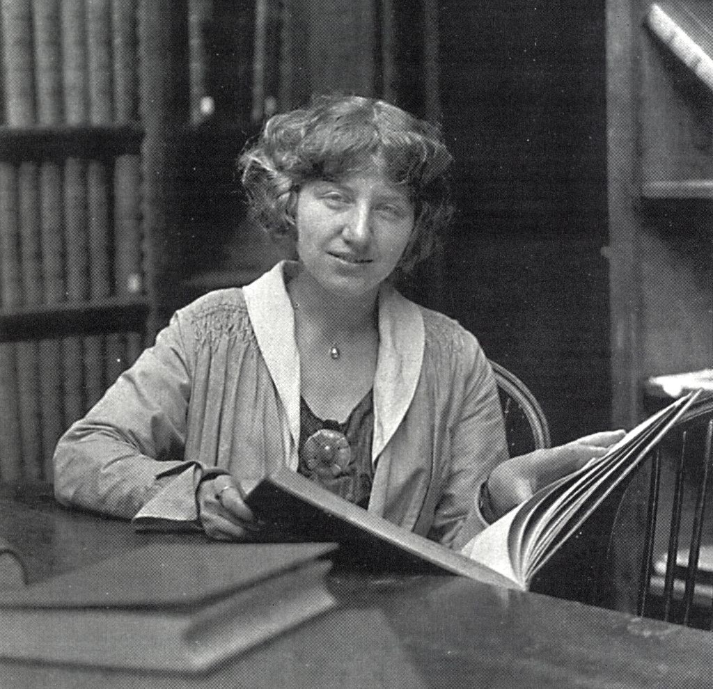 A vintage black and white image of architect Lutah Maria Riggs sitting behind a desk with a large book in her hands and bookcases behind her.